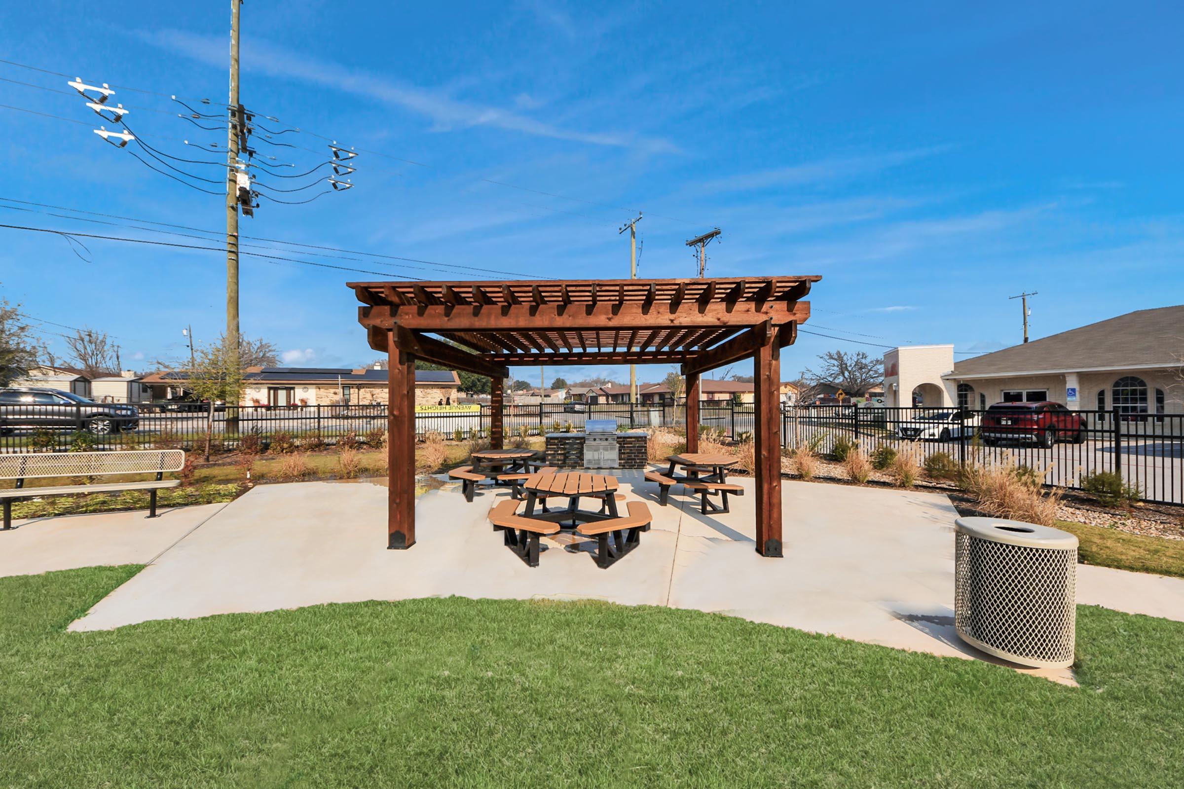 A shaded picnic area with a wooden pergola and multiple picnic tables on a grassy lawn. The background features a clear blue sky, utility poles, and a trash can. The setting is inviting for outdoor gatherings or meals.