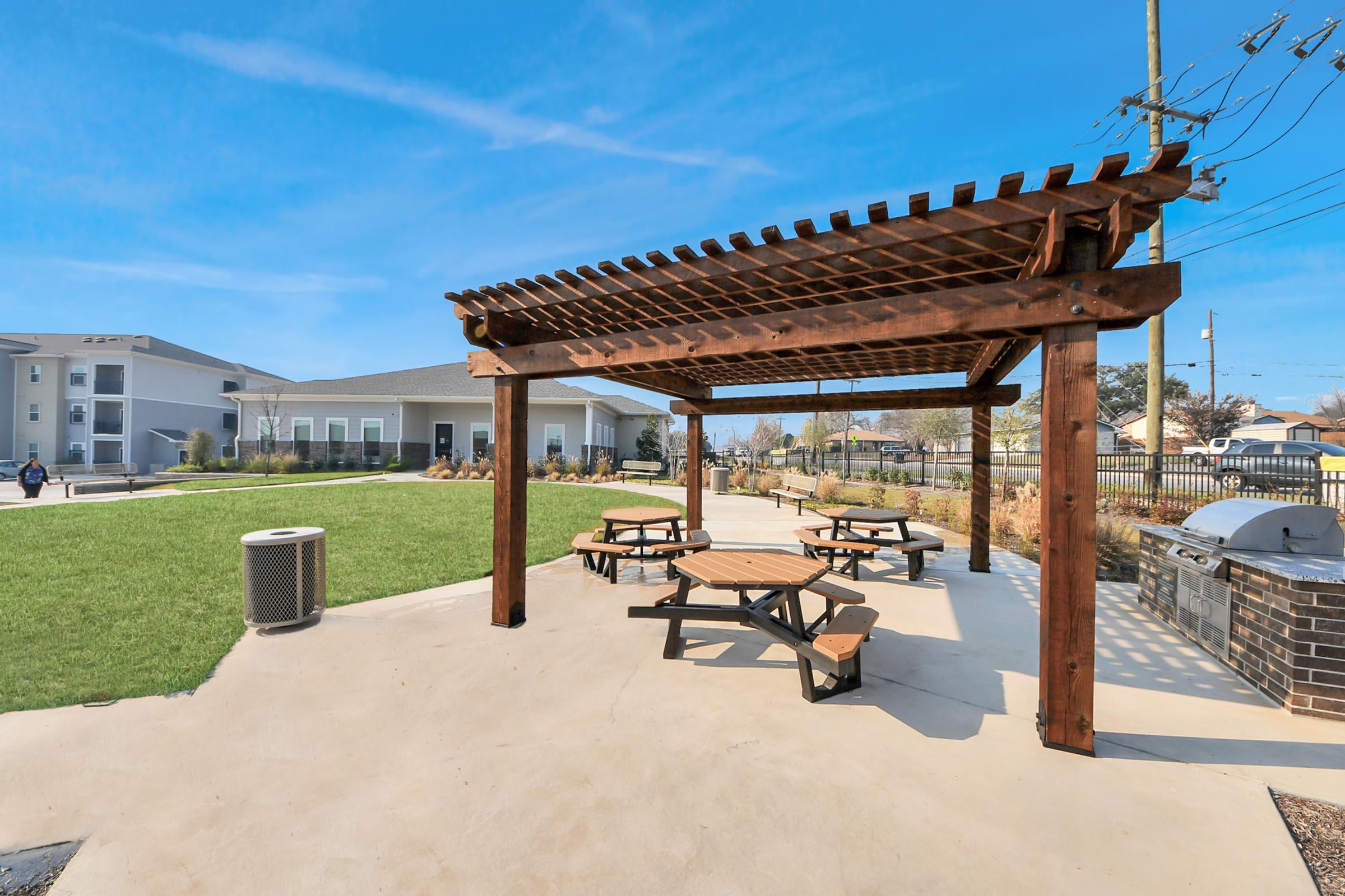 A shaded picnic area with wooden tables under a pergola, surrounded by grassy lawns and modern buildings in the background. Utility poles are visible, and a grilling station is nearby, creating a welcoming outdoor space for social gatherings.