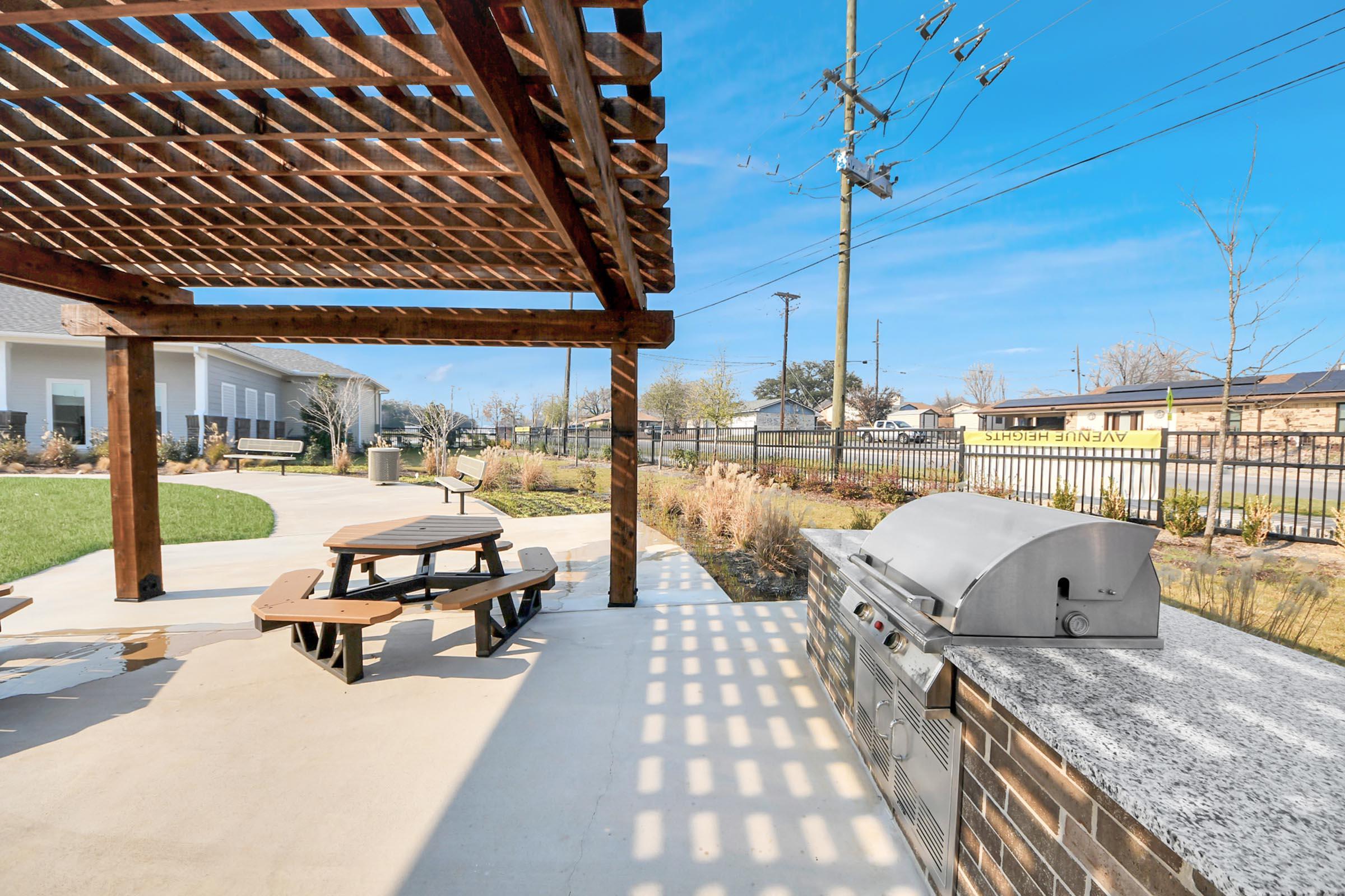 A shaded outdoor area with a picnic table and a built-in barbecue grill. The space features landscaped greenery and a clear blue sky. Nearby, power lines are visible, and a fence surrounds the area. The setting is ideal for gatherings and cooking outdoors.
