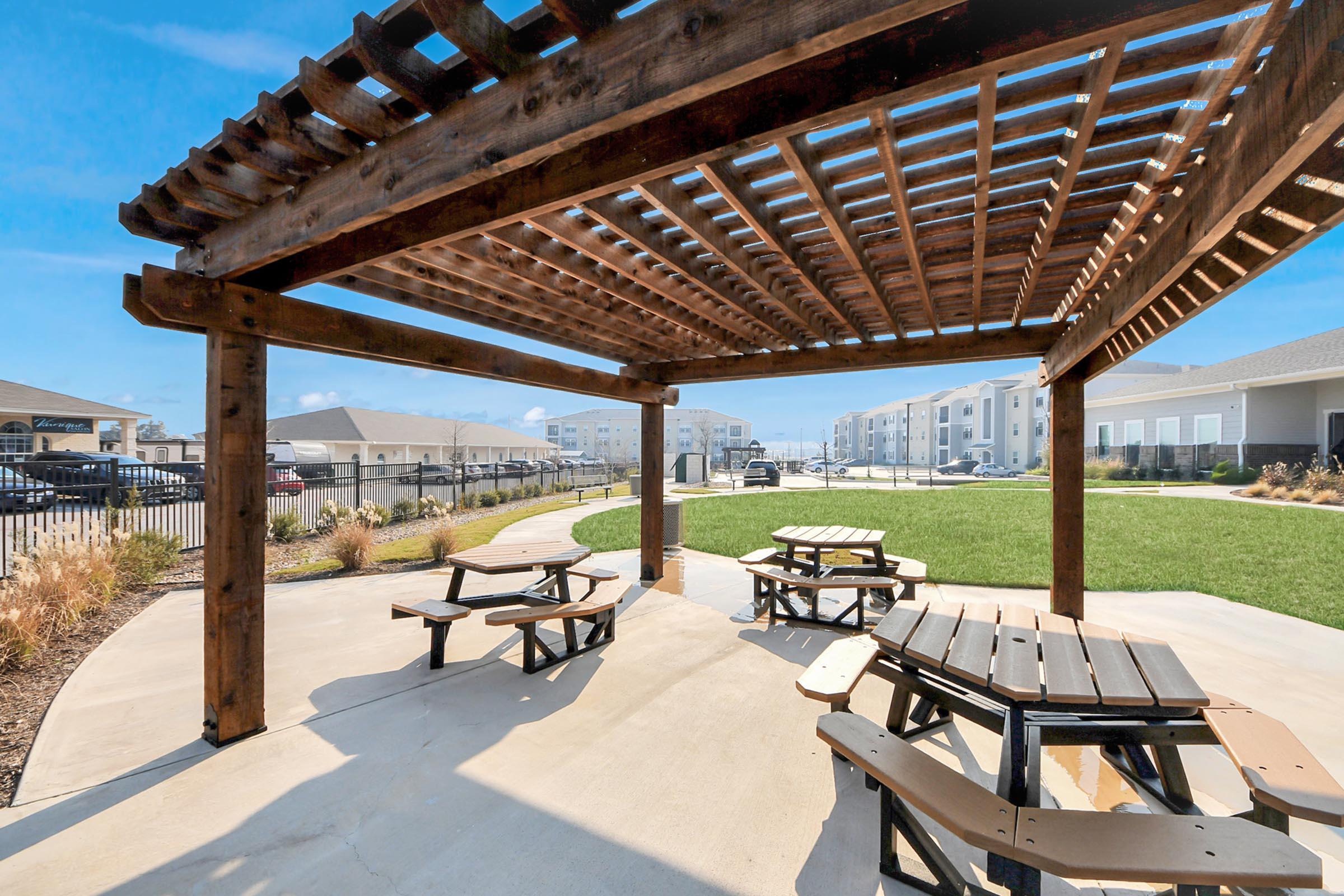 A shaded outdoor patio area with wooden picnic tables underneath a pergola. The ground is paved, and in the background, there are residential buildings surrounded by grassy areas. Clear blue skies are visible, creating a bright and inviting atmosphere.