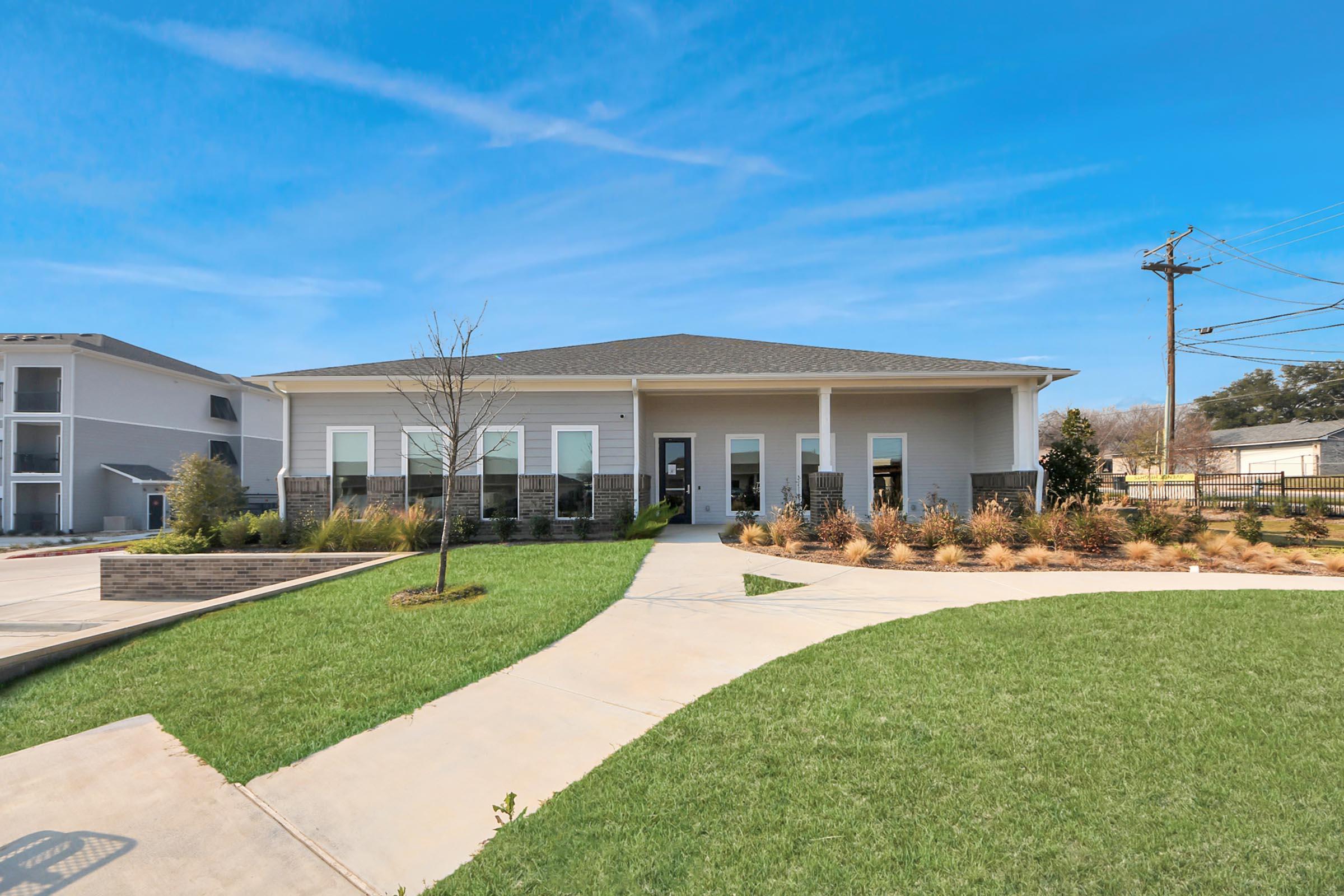 A modern single-story building with a sloped roof, surrounded by manicured lawns and pathways. There are shrubs and small trees in the landscaping, and the sky is clear with a few clouds. Utility poles are visible in the background, suggesting a residential or commercial area.