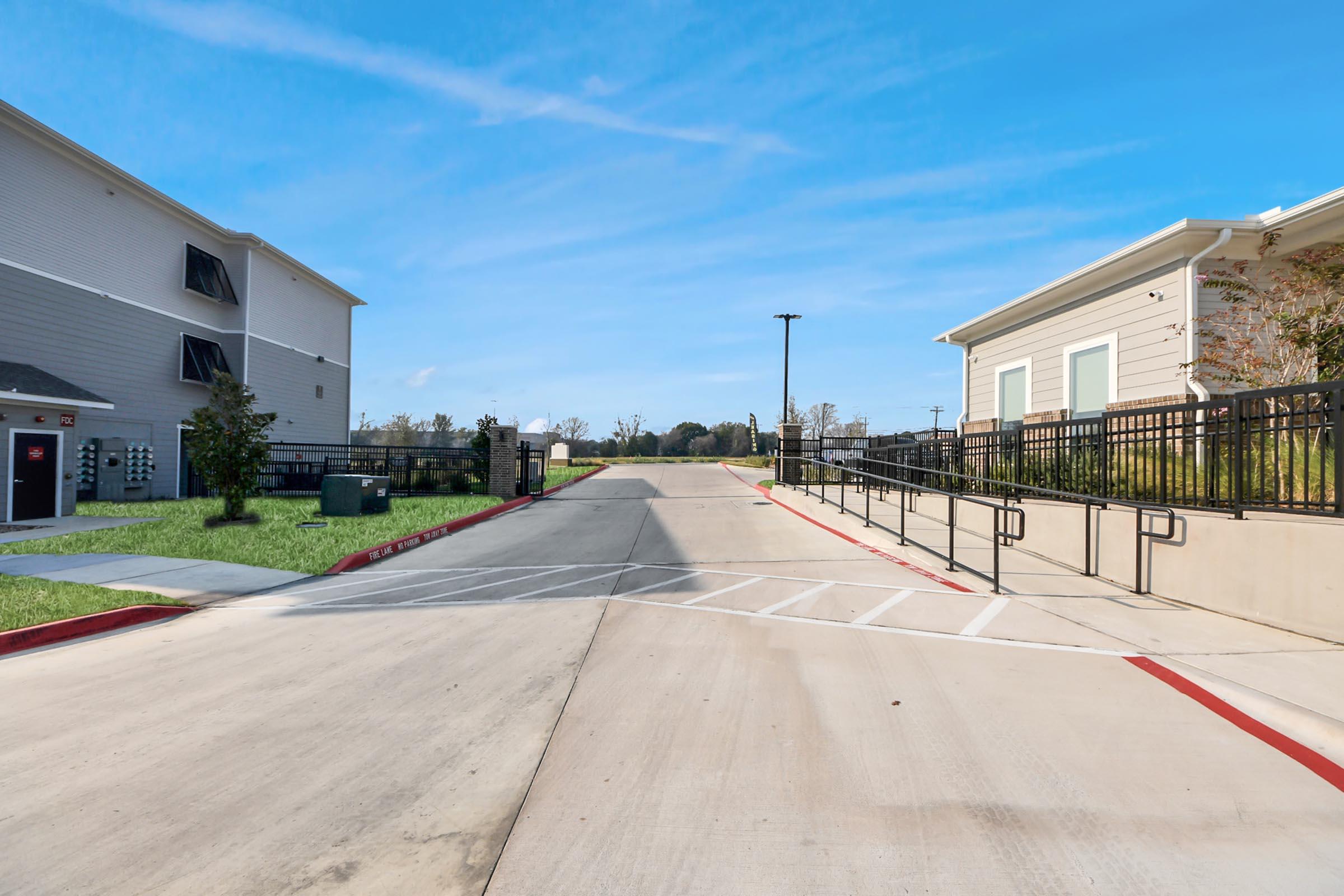 A view of a wide, paved pathway between two buildings, with manicured grass on either side. The sky is clear and blue, and there are railings along the pathway leading towards a distant area lined with trees. The scene suggests a well-maintained residential or commercial property.