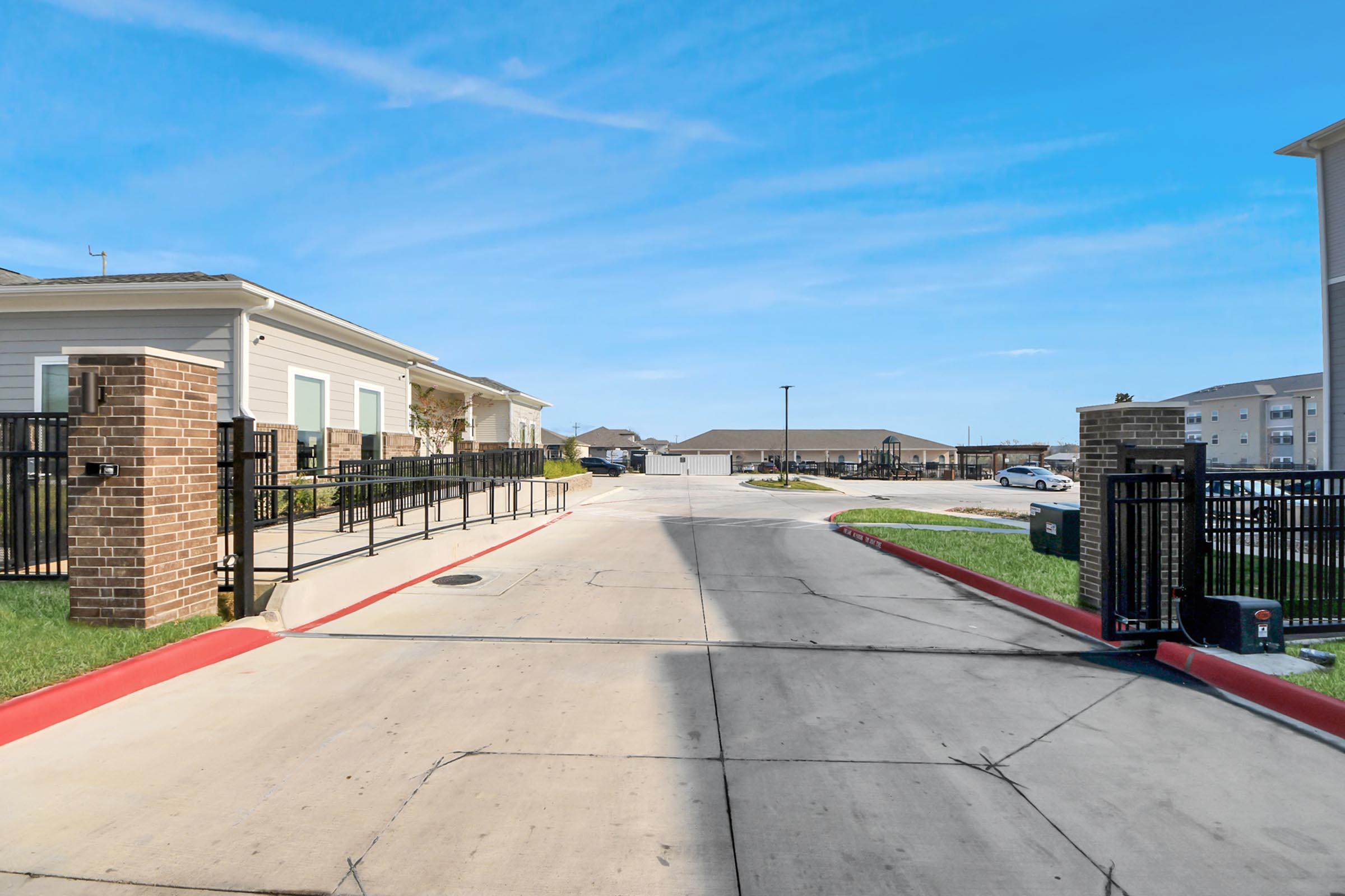 A view of a residential community entrance featuring a gated access point. The road is paved with light-colored asphalt, bordered by manicured grass. In the distance, multiple buildings are visible, suggesting a well-maintained neighborhood. The sky is clear and blue, enhancing the welcoming atmosphere.