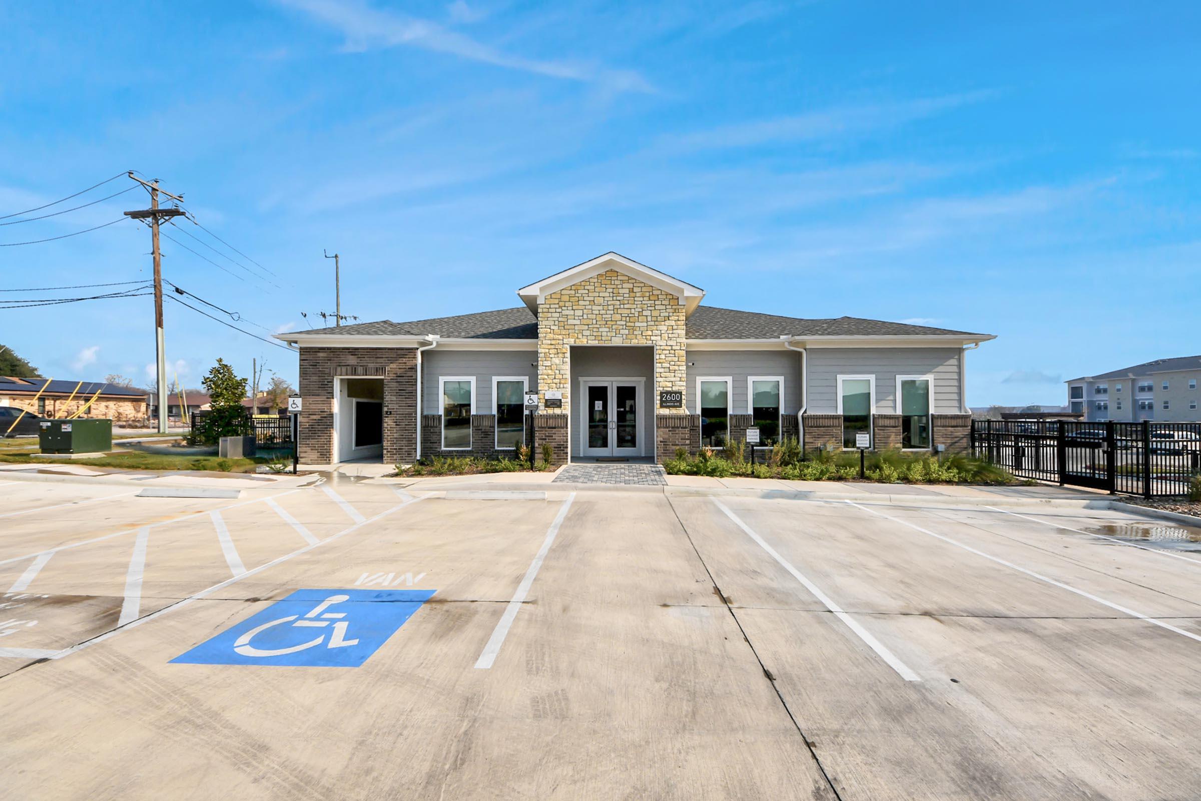 Single-story building with a stone and siding exterior, featuring multiple windows and a central entrance. In front, there is a parking area with a prominent blue handicap-accessible parking space. The surrounding area is paved, and the sky is clear with a few clouds.