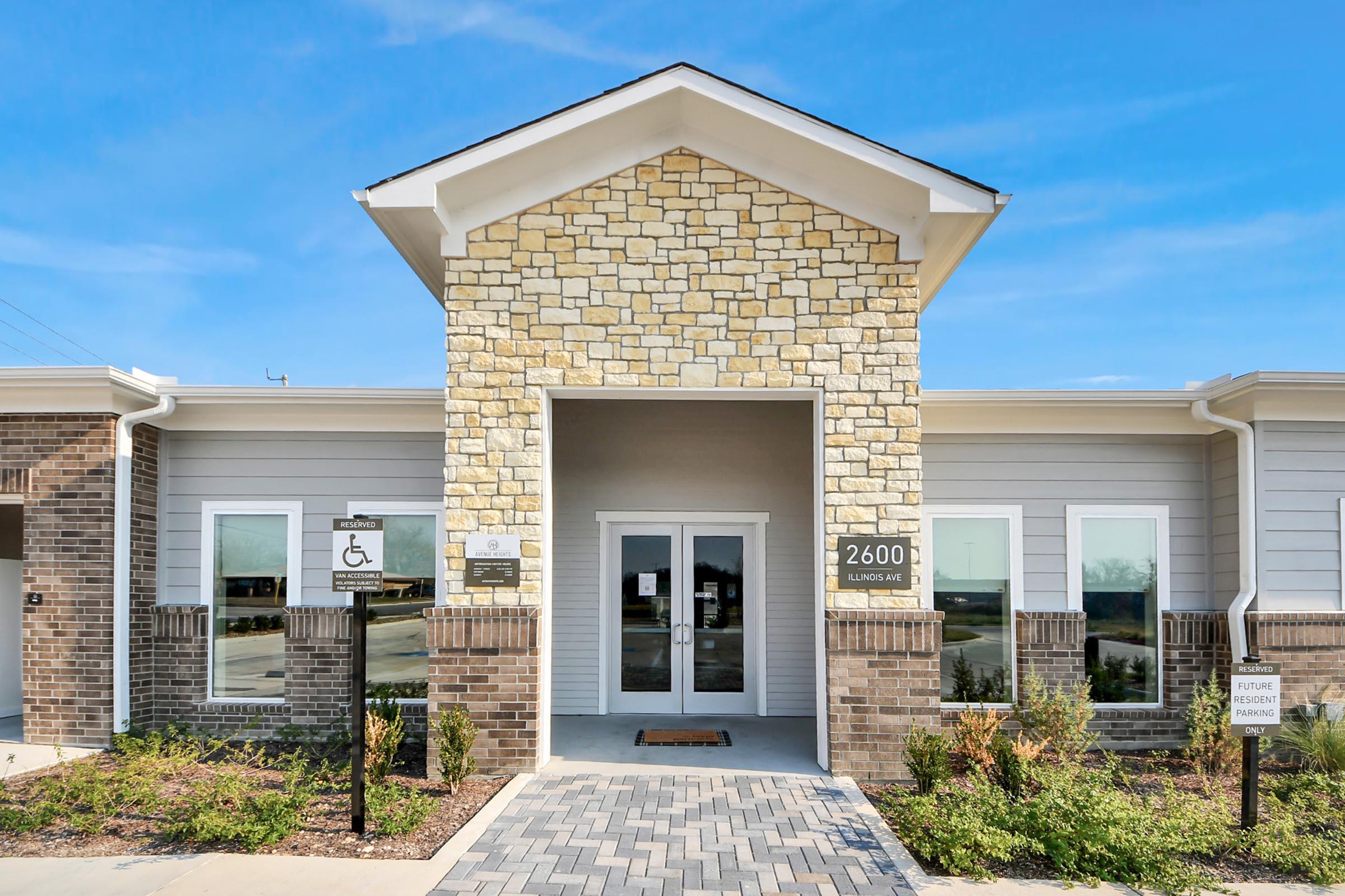 Exterior view of a modern building featuring a stone and brick facade. The entrance has double glass doors with a welcome mat, flanked by disability access signage. The address "2600" is displayed prominently above the entrance. Surrounding the entrance are landscaped areas with plants and clear blue skies in the background.