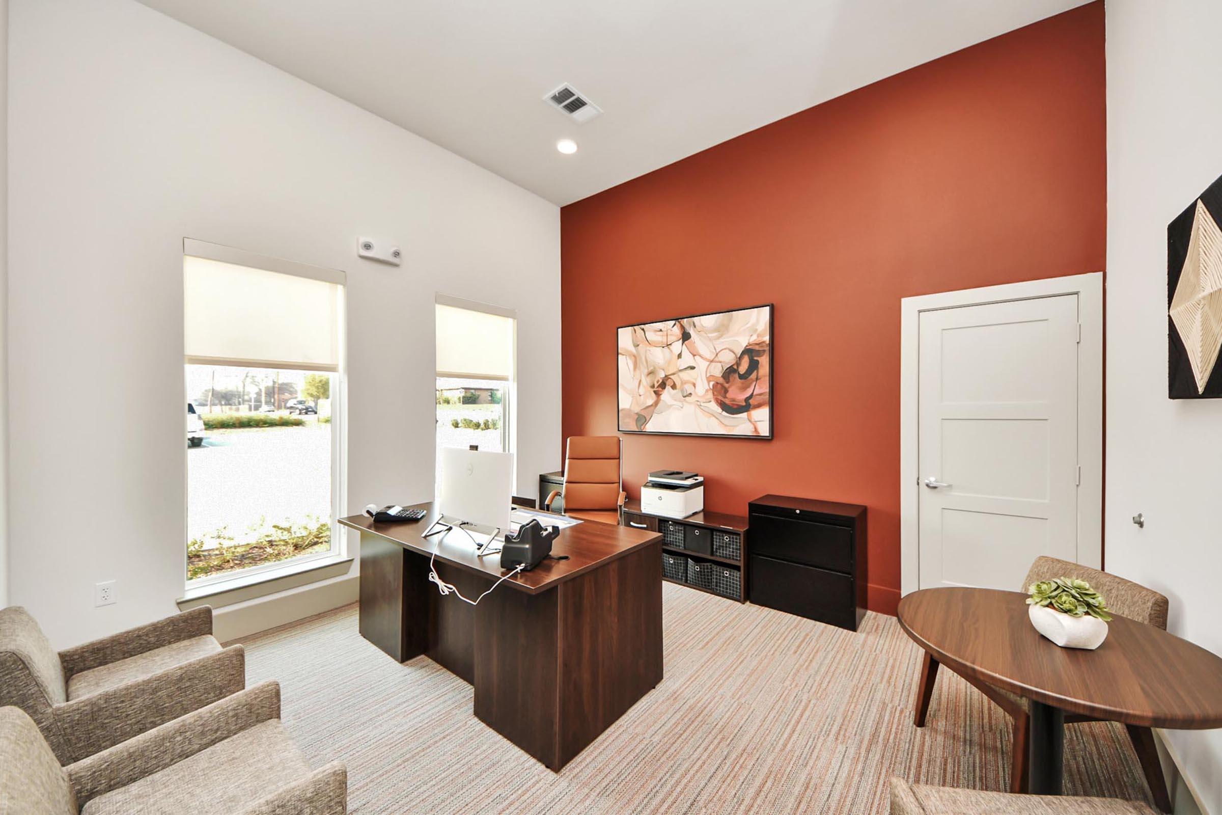 A modern office space featuring a wooden desk with a computer, printer, and office supplies. There are two beige chairs and a round table with a small plant. The walls are a mix of white and a warm orange, with large windows that provide natural light. A piece of abstract art is hung on the wall.