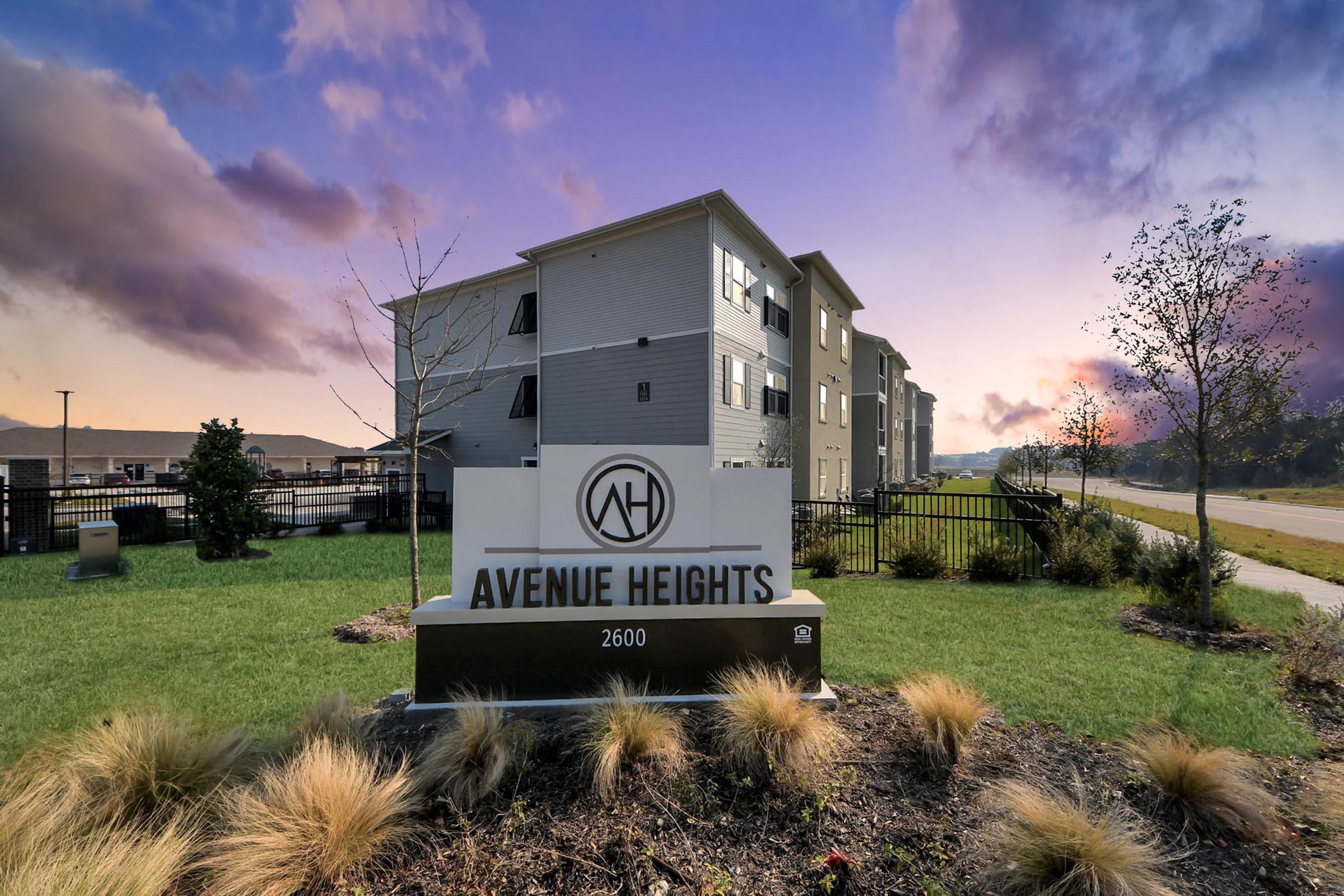 Avenue Heights apartment complex entrance sign featuring the logo, located at 2600. Surrounding greenery and landscaping with decorative plants are visible, alongside a road and residential buildings in the background under a colorful sky.