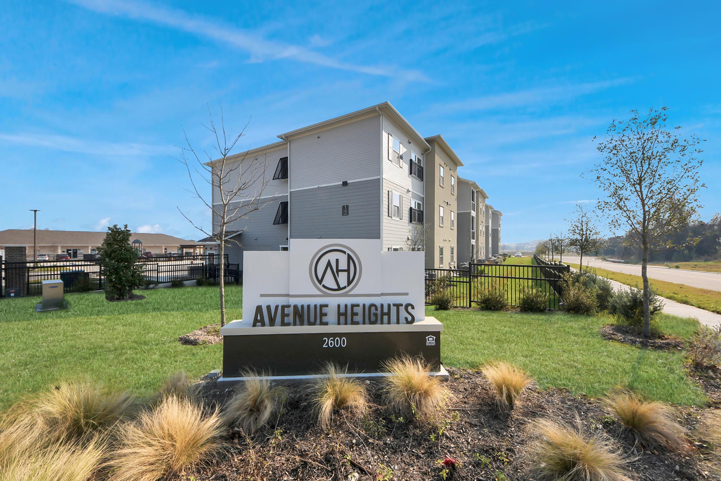 Sign for Avenue Heights, featuring the name prominently. The setting includes a well-maintained landscape with grass and ornamental plants, along with a multi-story residential building in the background. A road runs beside the property, under a clear blue sky.