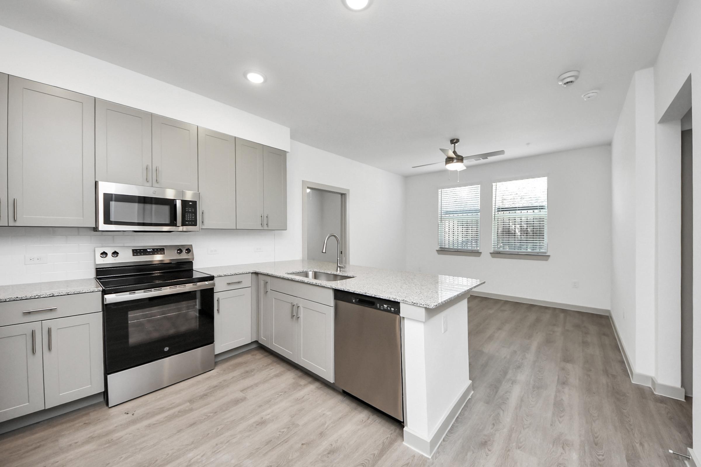 A modern kitchen with gray cabinets, stainless steel appliances, a granite countertop, and an open layout. It features a microwave, stove, sink, and dishwasher, with a light and airy living space visible in the background, highlighted by natural light from two windows.