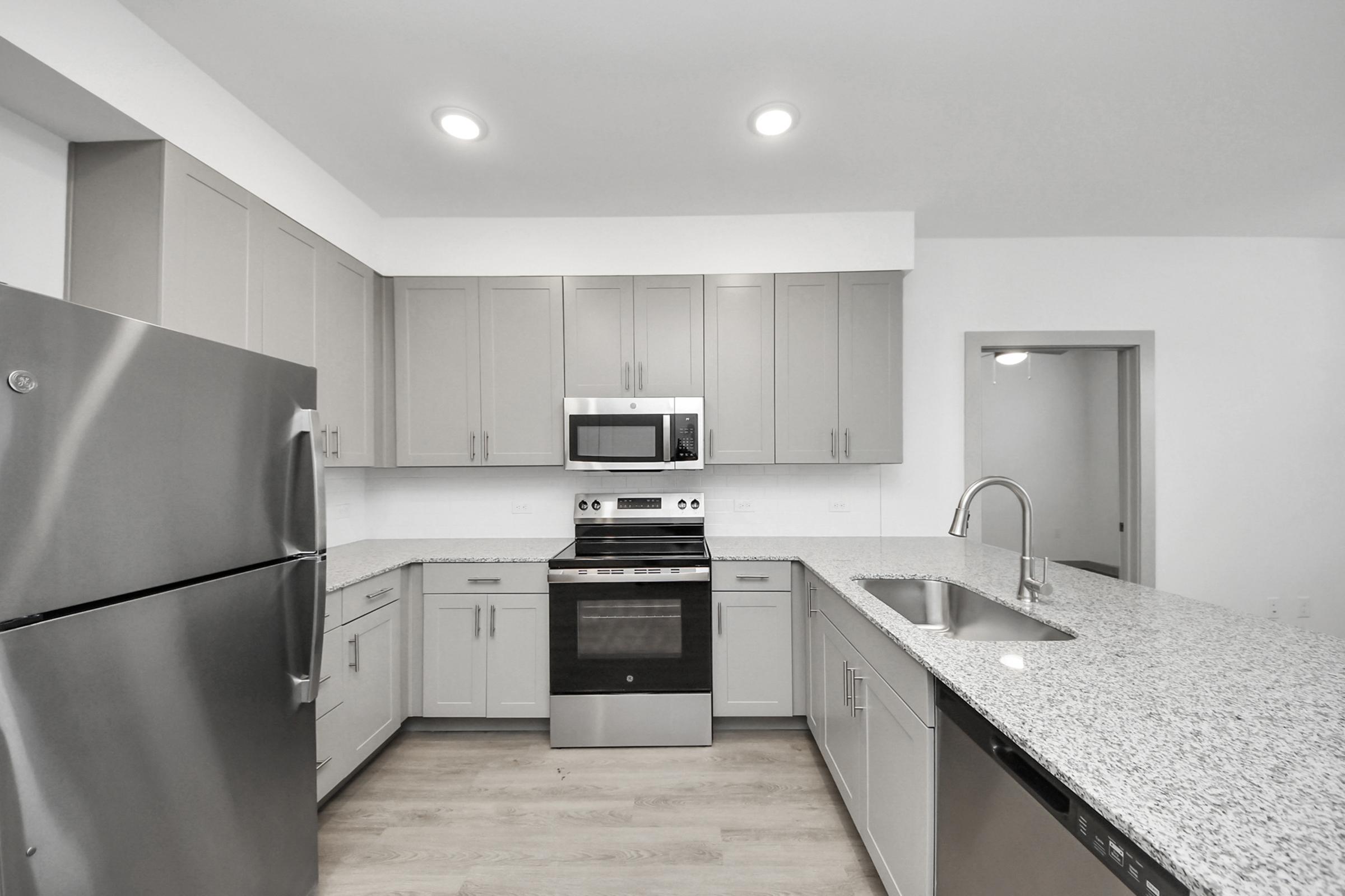 Modern kitchen featuring stainless steel appliances, including a refrigerator, oven, and microwave. The cabinets are light gray, and the countertop is speckled granite. A sleek sink is integrated into the island, which offers additional counter space. The kitchen has bright overhead lighting and a minimalist design.