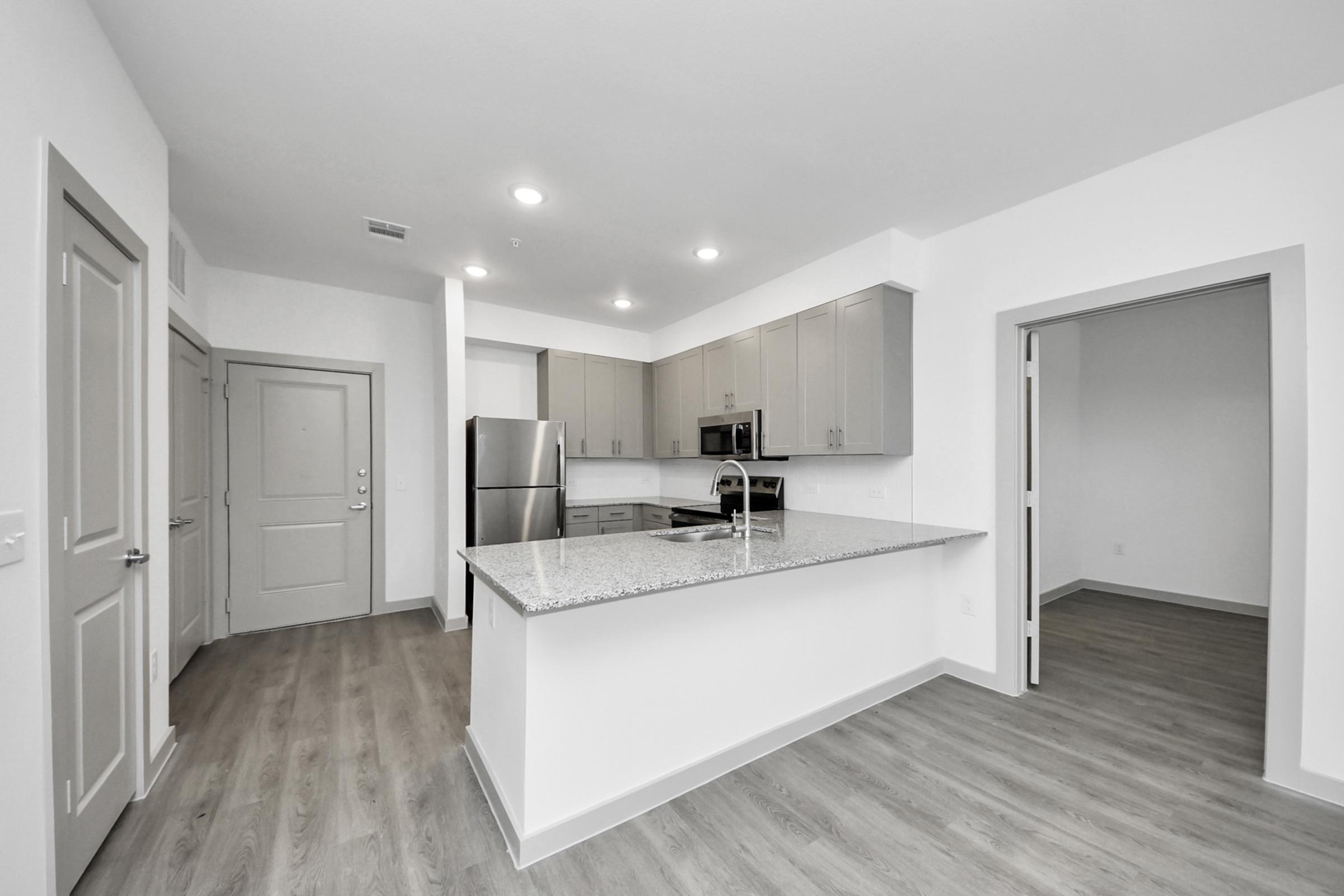 Modern kitchen in a spacious apartment featuring light-colored cabinets, stainless steel appliances, and a large granite island. The open floor plan includes a view into a living area with hardwood-like flooring and access to a well-lit entryway. Bright, minimalist design with neutral tones.