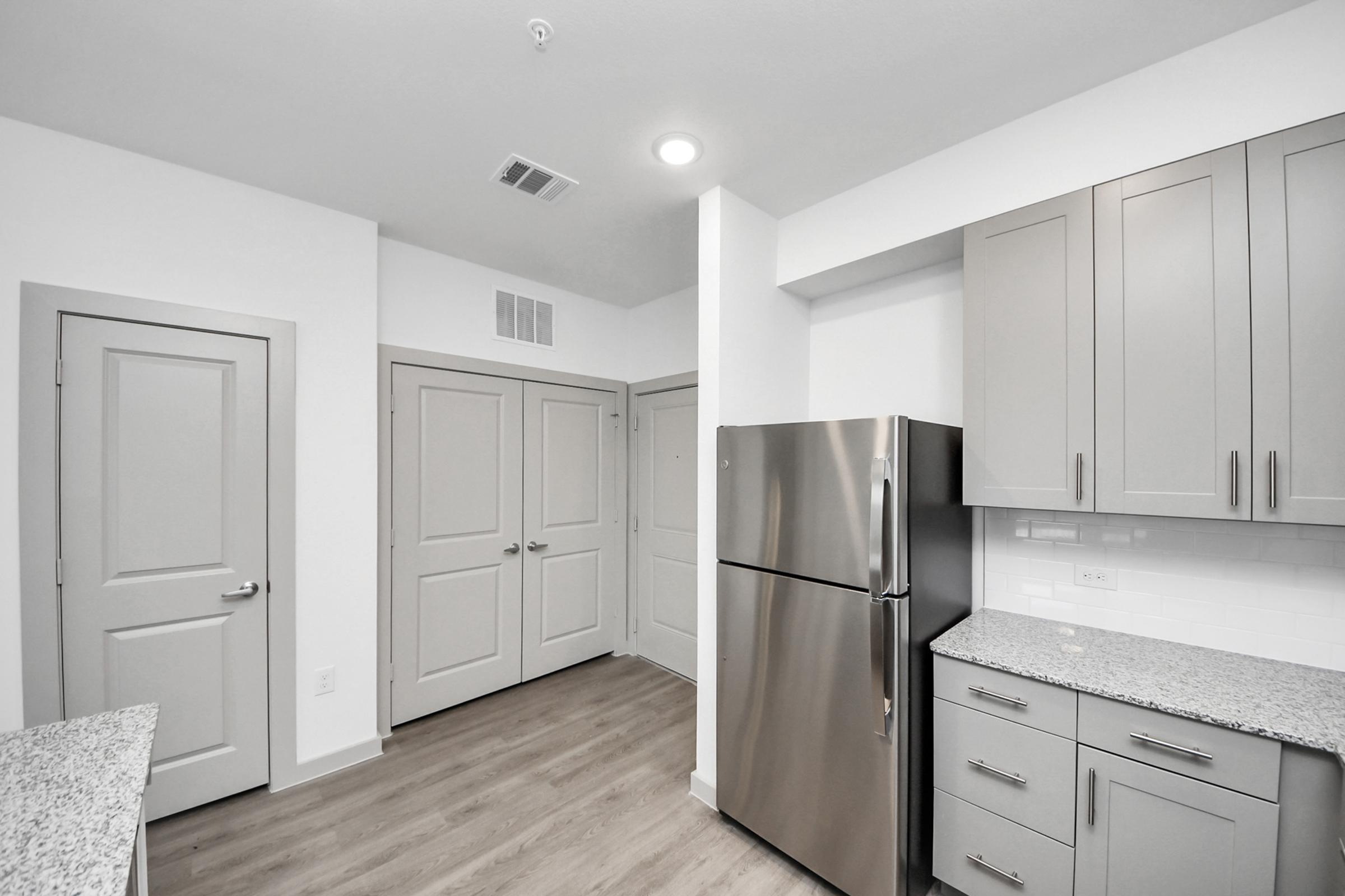 Modern kitchen featuring stainless steel refrigerator, light gray cabinets, and granite countertops. The space includes a doorway leading to additional rooms, with light-colored walls and wood-like flooring, creating a bright and airy atmosphere.