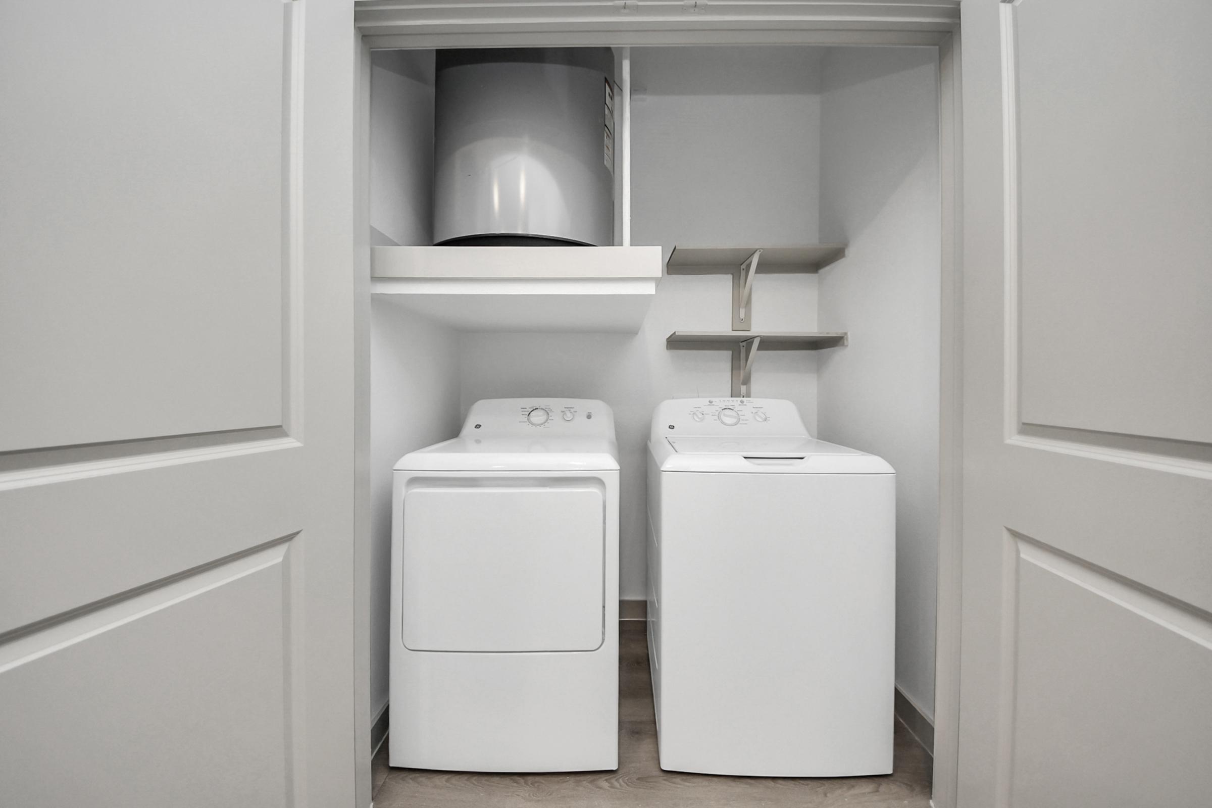 A modern laundry room featuring a stacked washer and dryer in a white finish. The appliances are neatly arranged in a closet space with light-colored shelving above for storage. The room has light wooden flooring and smooth, neutral-colored walls, creating a clean and organized appearance.