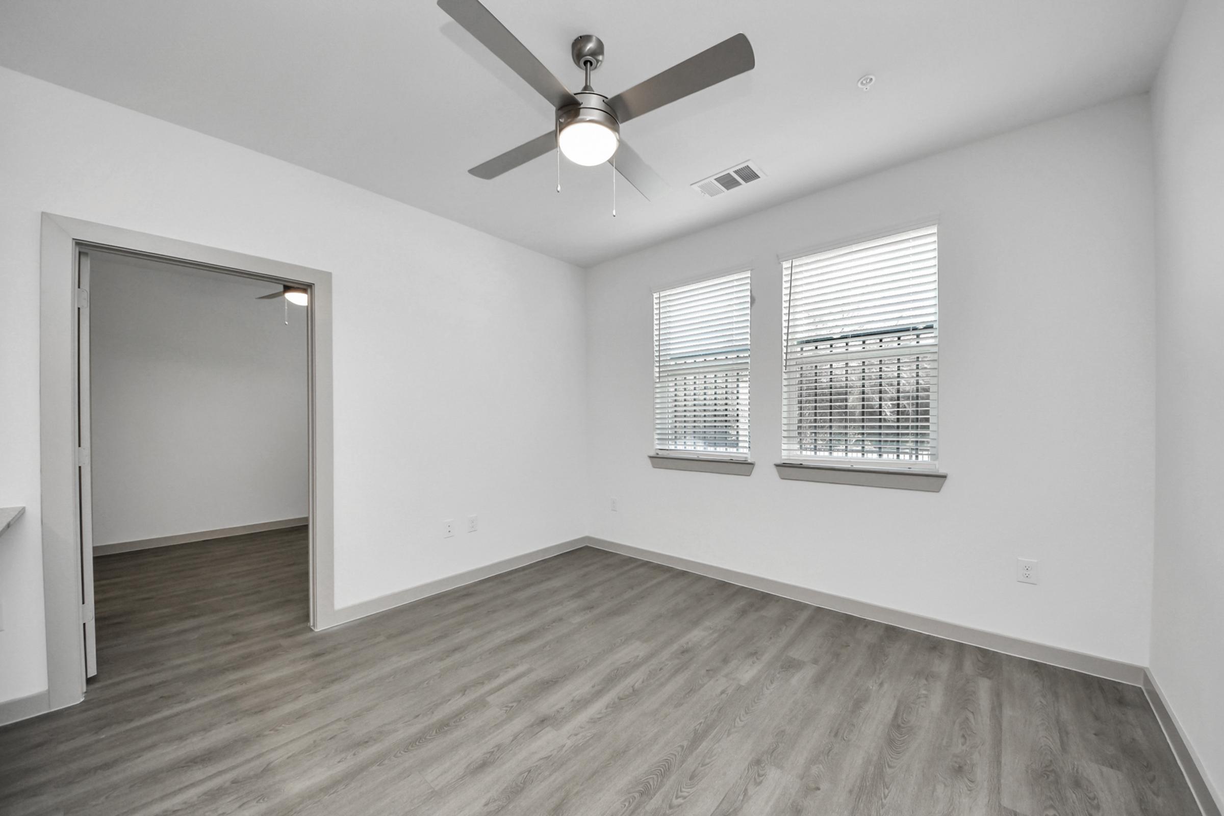 A spacious, modern room with light gray walls and timber-style flooring. It features a ceiling fan, two windows with blinds allowing natural light, and a doorway leading to another room. The minimalist design emphasizes openness and allows for flexible use of space.