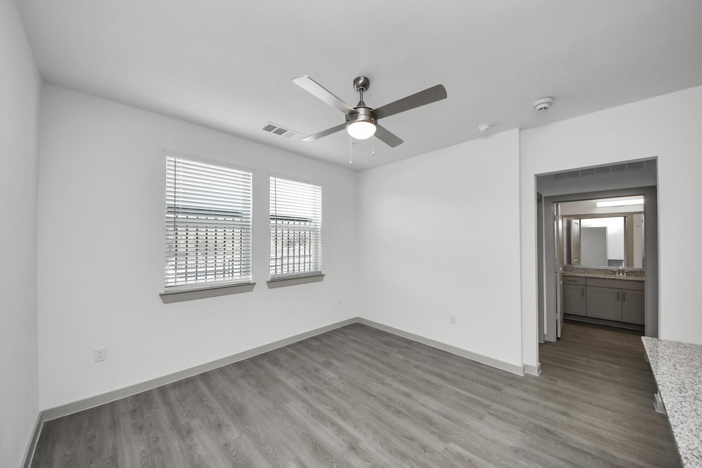 Bright and airy interior of a modern room featuring light gray walls, large windows with white blinds, and a ceiling fan. The space has laminate flooring and an adjacent room with a bathroom visible in the background. Clean and minimalistic design with ample natural light.