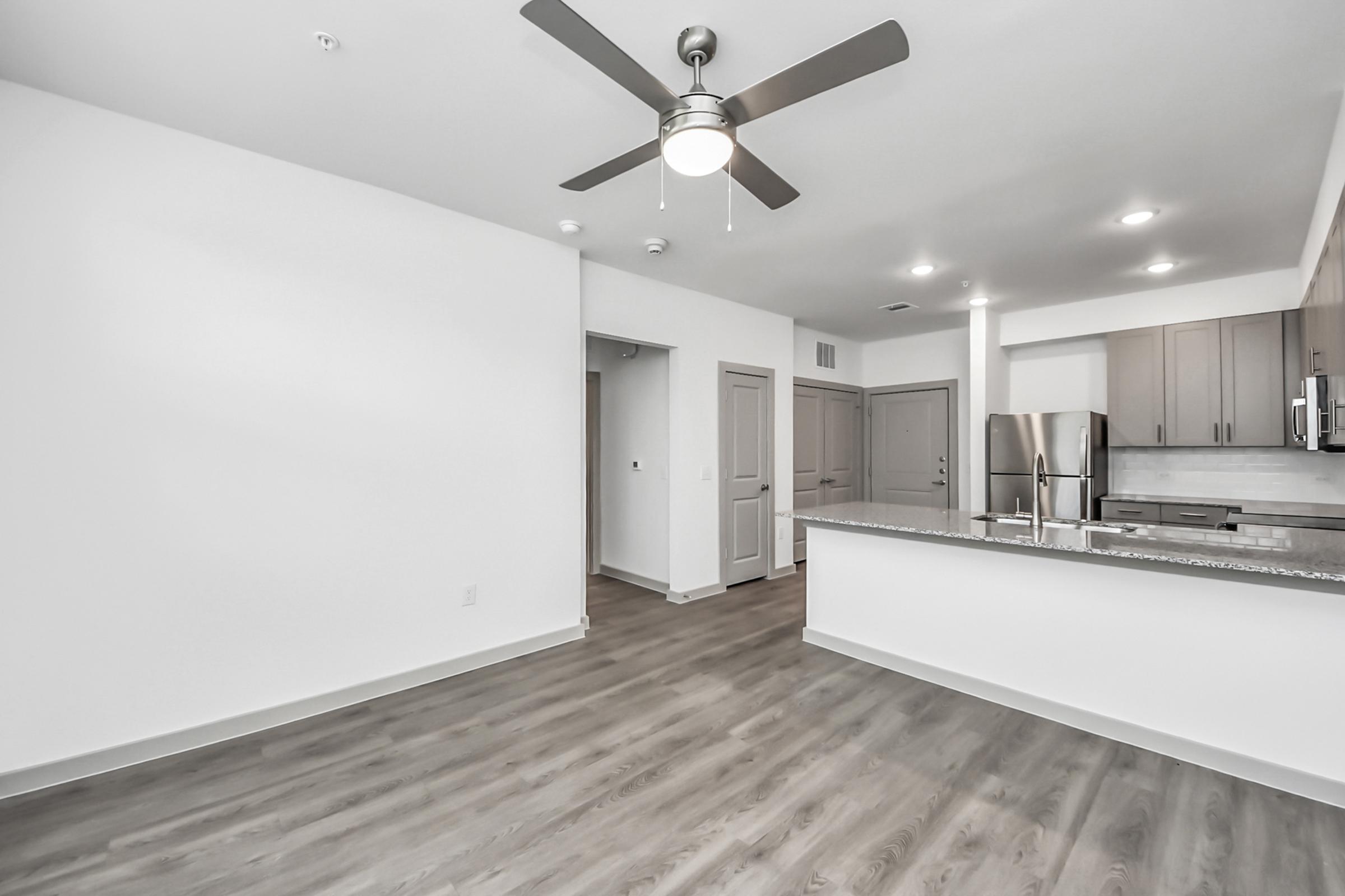 Interior view of a modern, open-concept living space featuring a kitchen with stainless steel appliances, a ceiling fan, and light-colored walls. The flooring is light wood, and there are doors leading to other rooms visible in the background. The area is bright and airy, suggesting a contemporary design.
