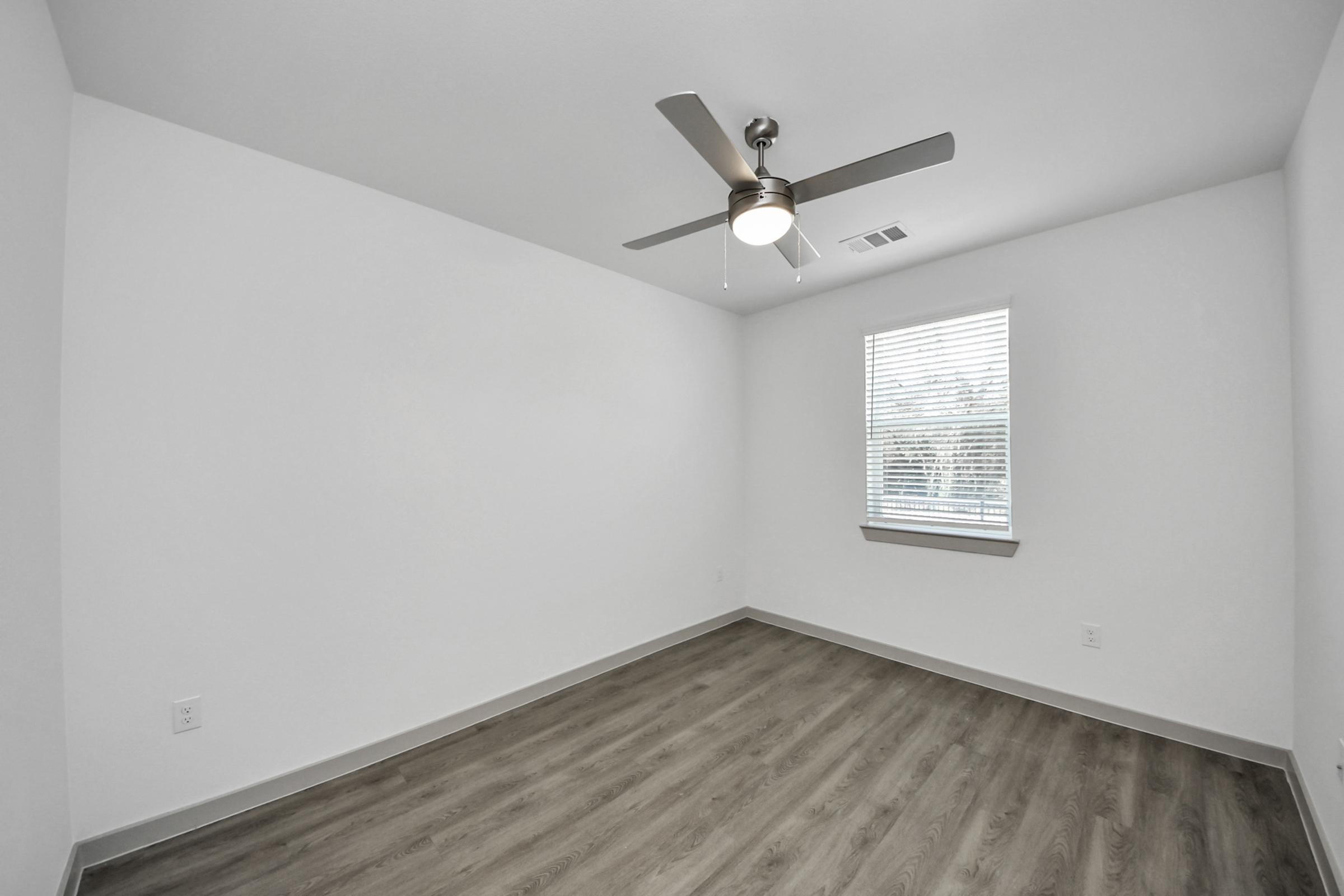 Empty room with light-colored walls and a ceiling fan. A window with blinds allows natural light in, and the floor features light wood or laminate. The overall space has a minimalist design, creating a bright and open atmosphere.