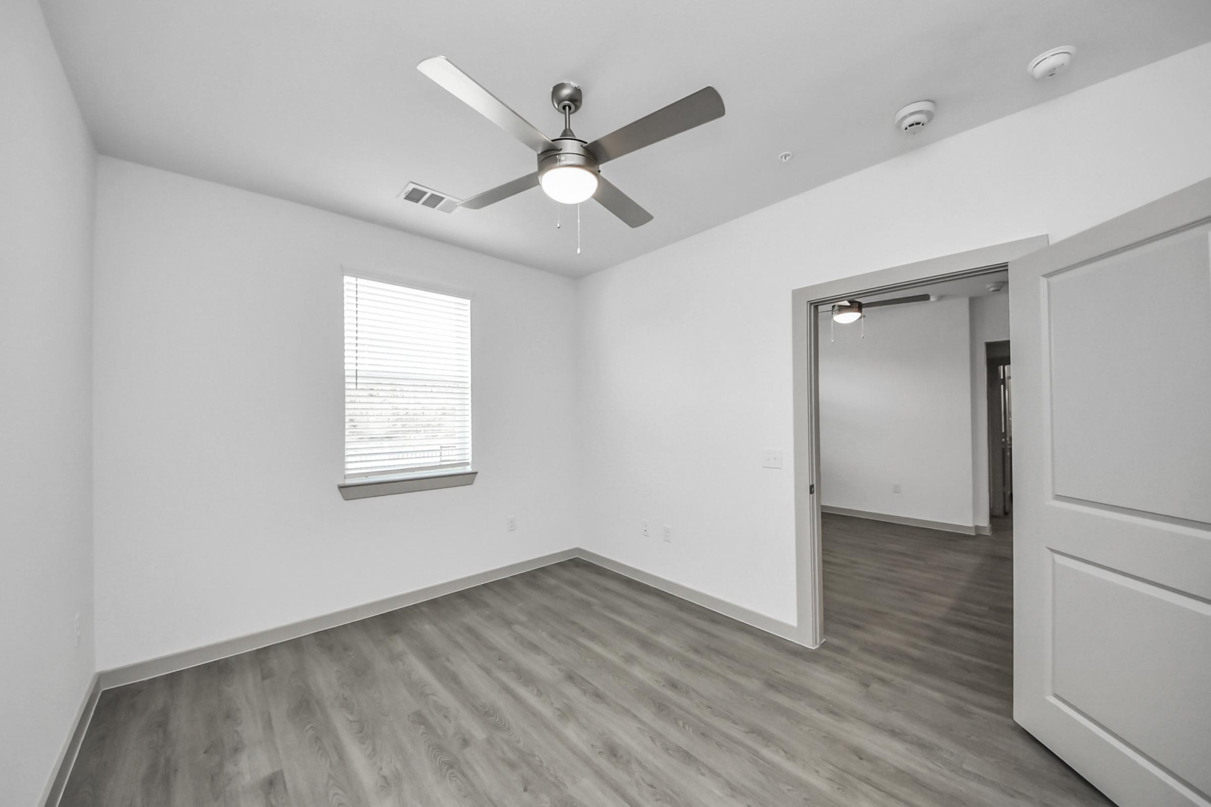 Interior view of a modern, empty room with light gray walls and wood-like flooring. A ceiling fan is mounted above, and a window with blinds allows natural light in. A door on the right leads to another area, enhancing the spacious feel of the room.