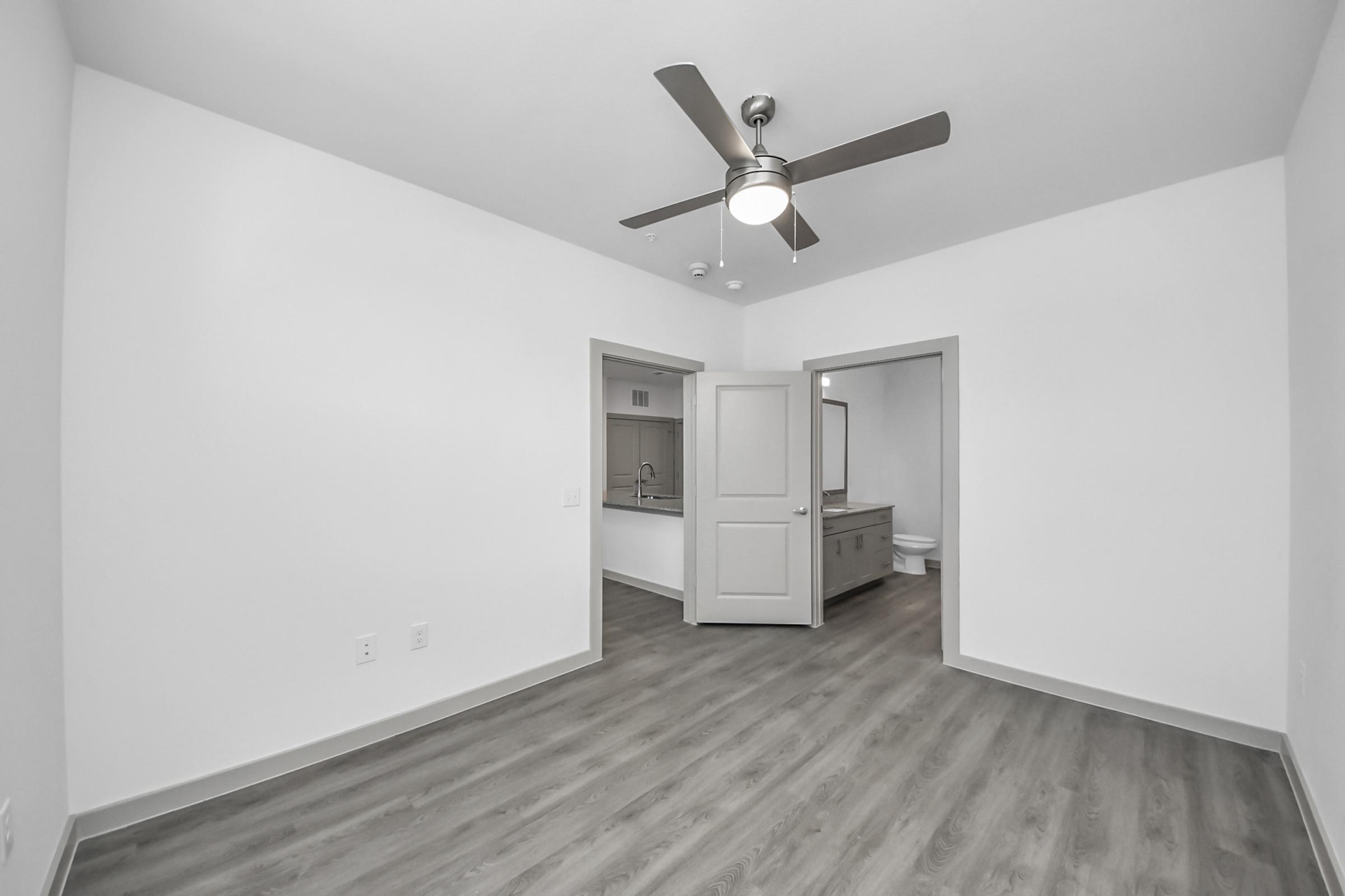 A spacious, empty room featuring a ceiling fan, light gray walls, and hardwood flooring. Two open doorways lead to an adjoining room and a bathroom. The overall aesthetic is modern and minimalist, with ample natural light and an open layout.