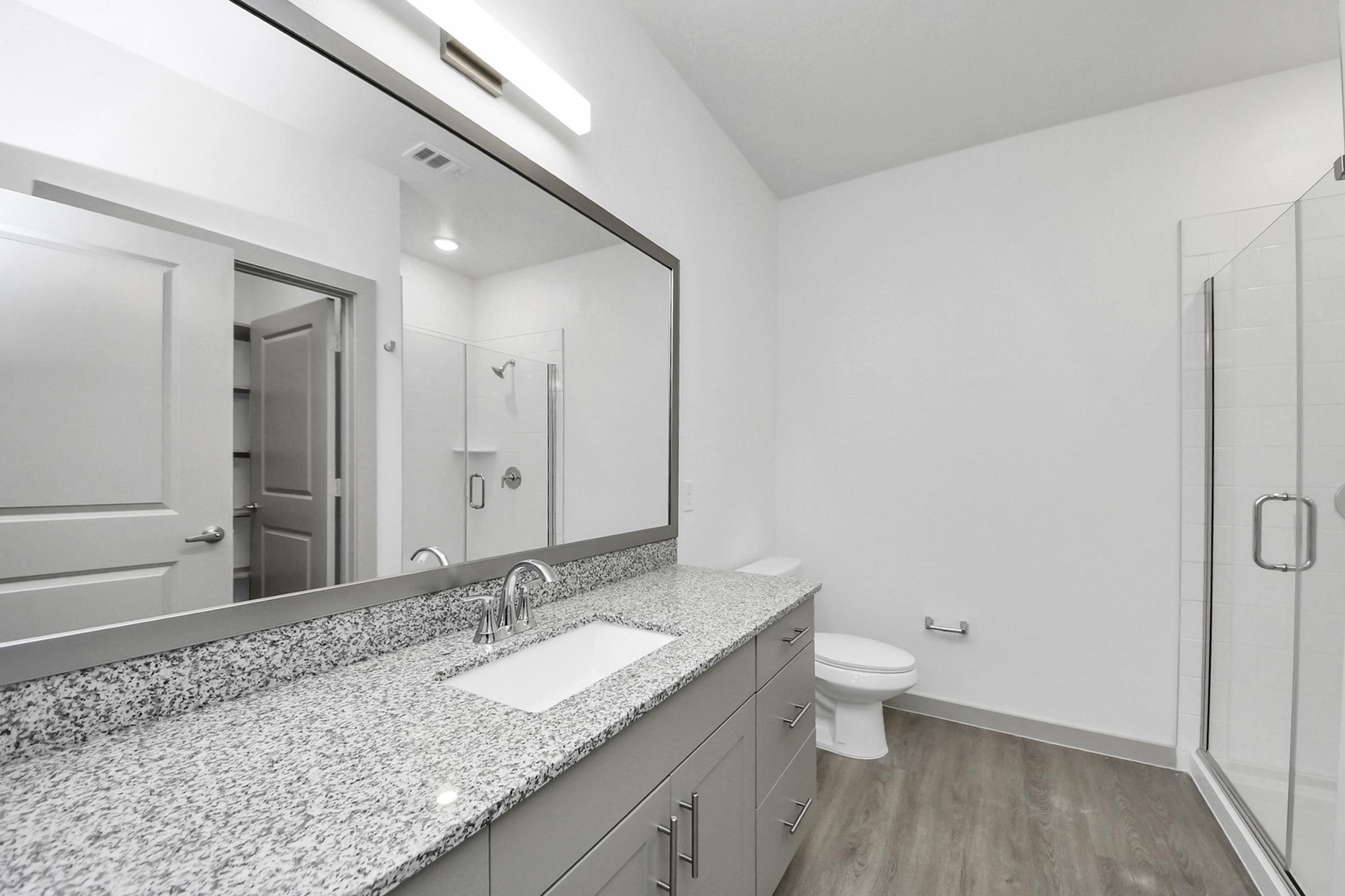 A modern bathroom featuring a large mirror above a granite countertop with a sink. The space includes a shower with glass doors, a toilet, and light-colored walls. A door leads to another room, adding to the sleek and contemporary design. Natural light illuminates the area.