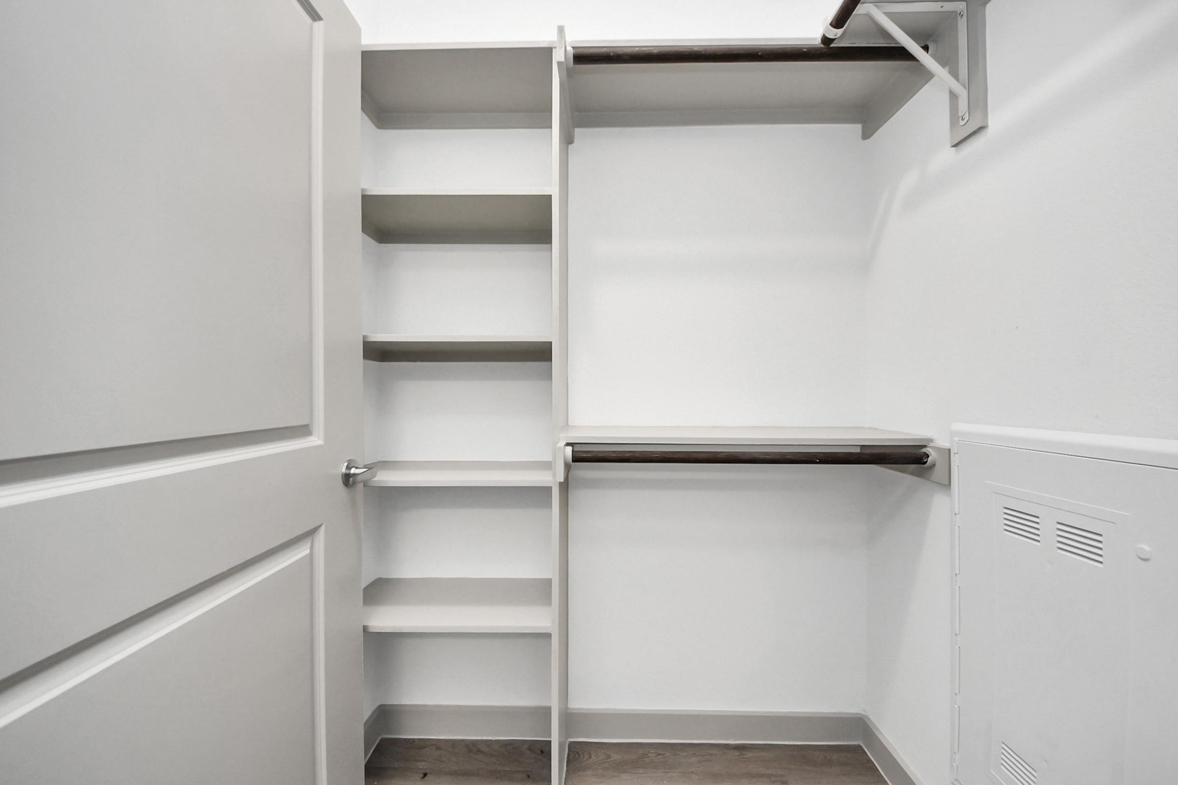 A clean and organized closet space featuring several open shelves, a hanging rod, and a small table-like surface. The walls are painted white, and the floor is a light-colored wood laminate. The door is closed and there are no items visible inside the closet.