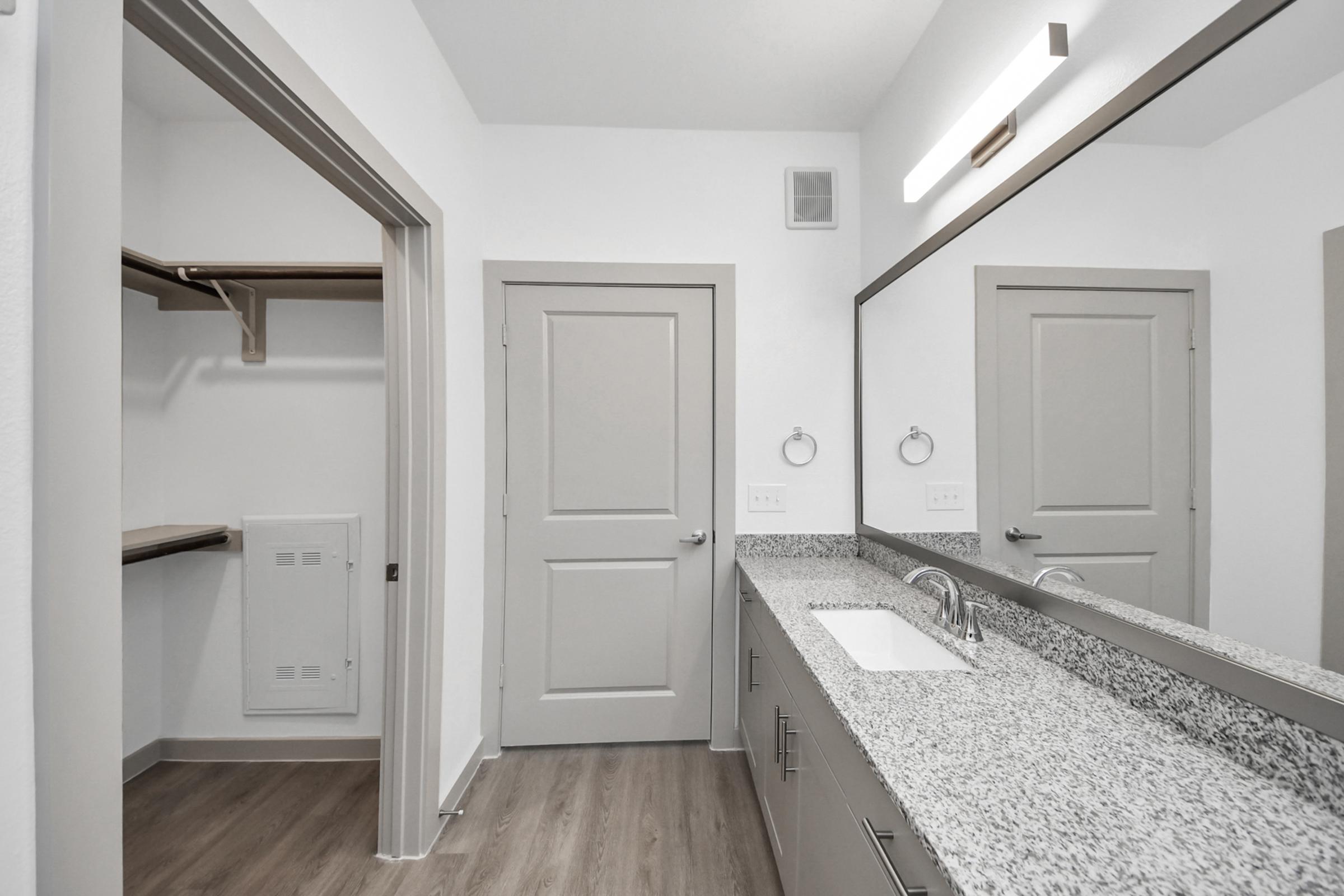 A modern bathroom featuring a double vanity with a granite countertop and mirror, gray cabinetry, and a silver wall-mounted light fixture. To the left, there’s an open closet space with hanging rods, complemented by a door leading to another room. Neutral-colored walls and wooden flooring enhance the spacious feel.