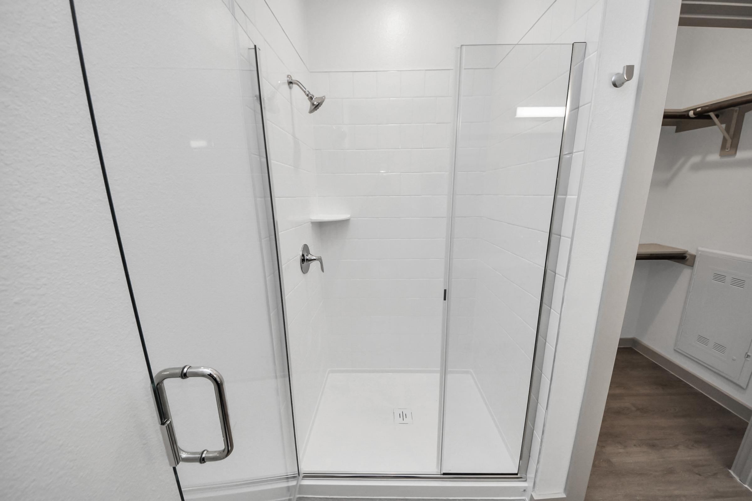 A clean, modern shower enclosure with clear glass doors, white tiled walls, and a built-in shelf. The shower features chrome fixtures, including a handheld showerhead and a wall-mounted showerhead. The floor is light-colored, and there's a small storage area visible nearby.