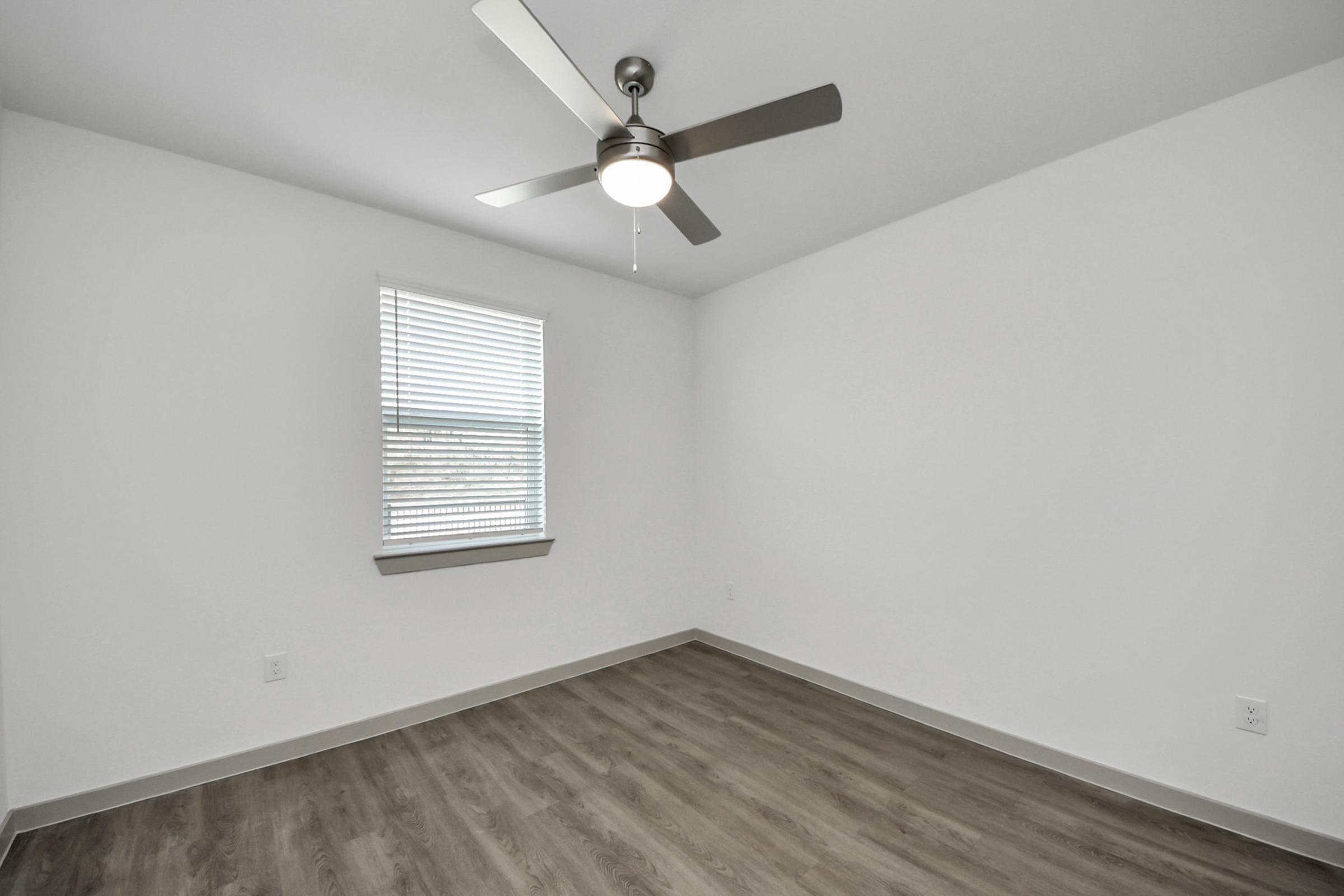 A minimalist room with light-colored walls and a ceiling fan. The space has a window with blinds allowing natural light, and the flooring is a warm wood-like laminate. There is ample open space, creating a clean and airy atmosphere.