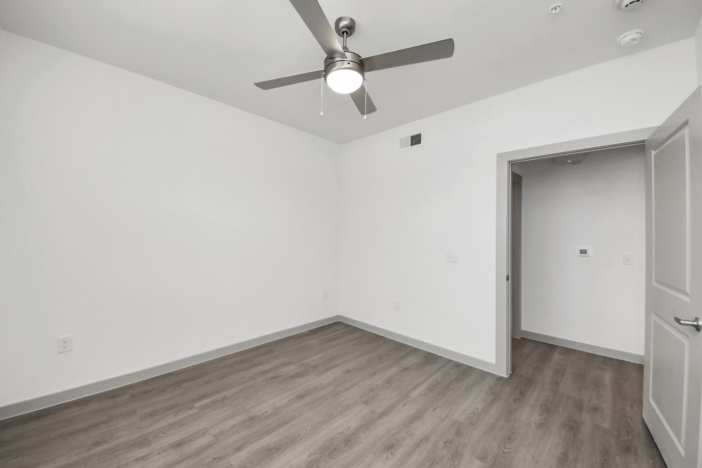 A spacious, empty room with light-colored walls and wooden flooring. It features a ceiling fan with a light fixture, a doorway leading to another space, and a wall-mounted thermostat. The room is bathed in natural light, creating a bright and airy atmosphere.