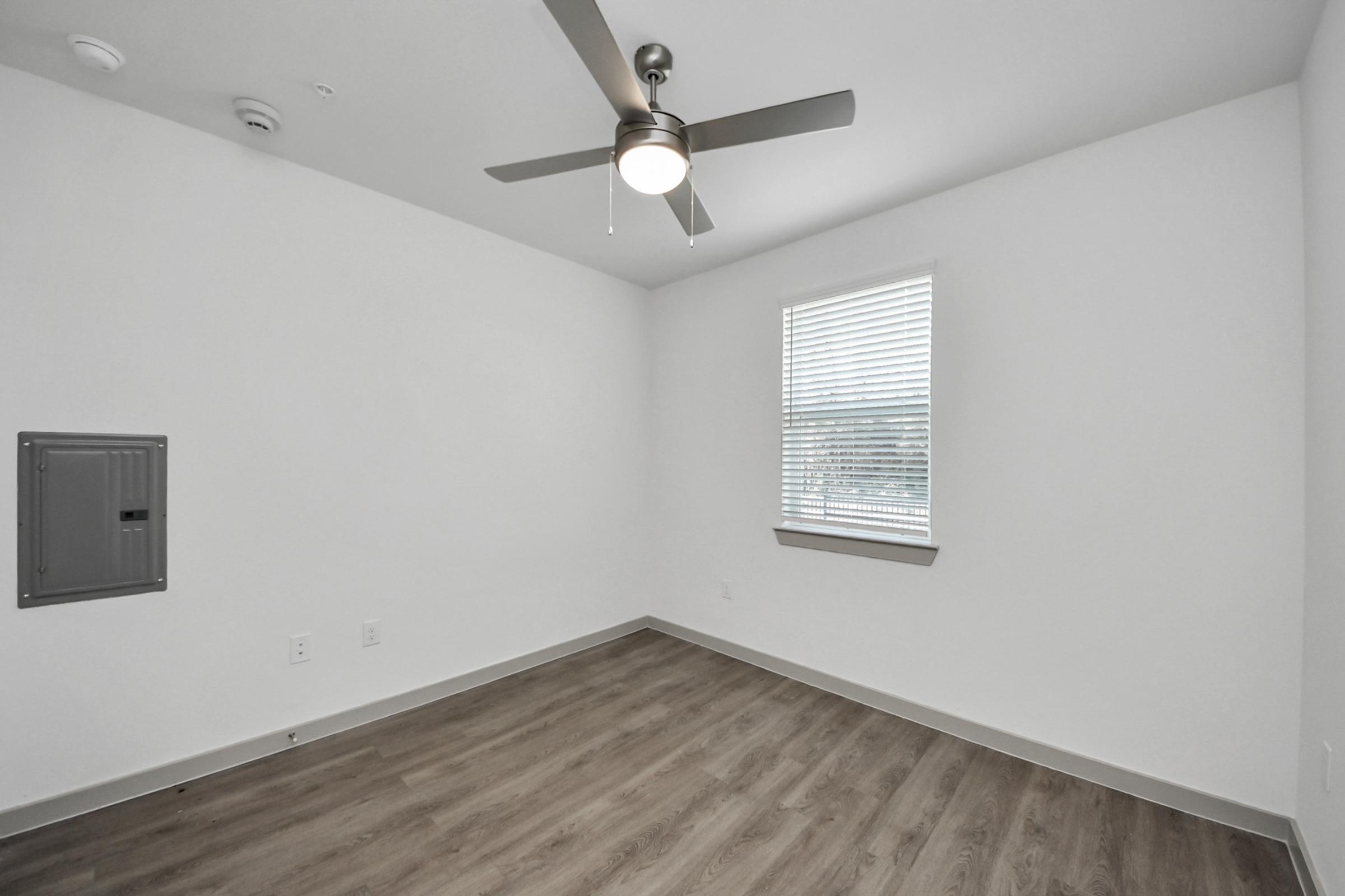 A small, empty room featuring a light-colored wall, brown laminate flooring, a ceiling fan, and a window with blinds. A wall-mounted electrical panel is visible on the left. The room has a modern and minimalist design, with natural light coming through the window.