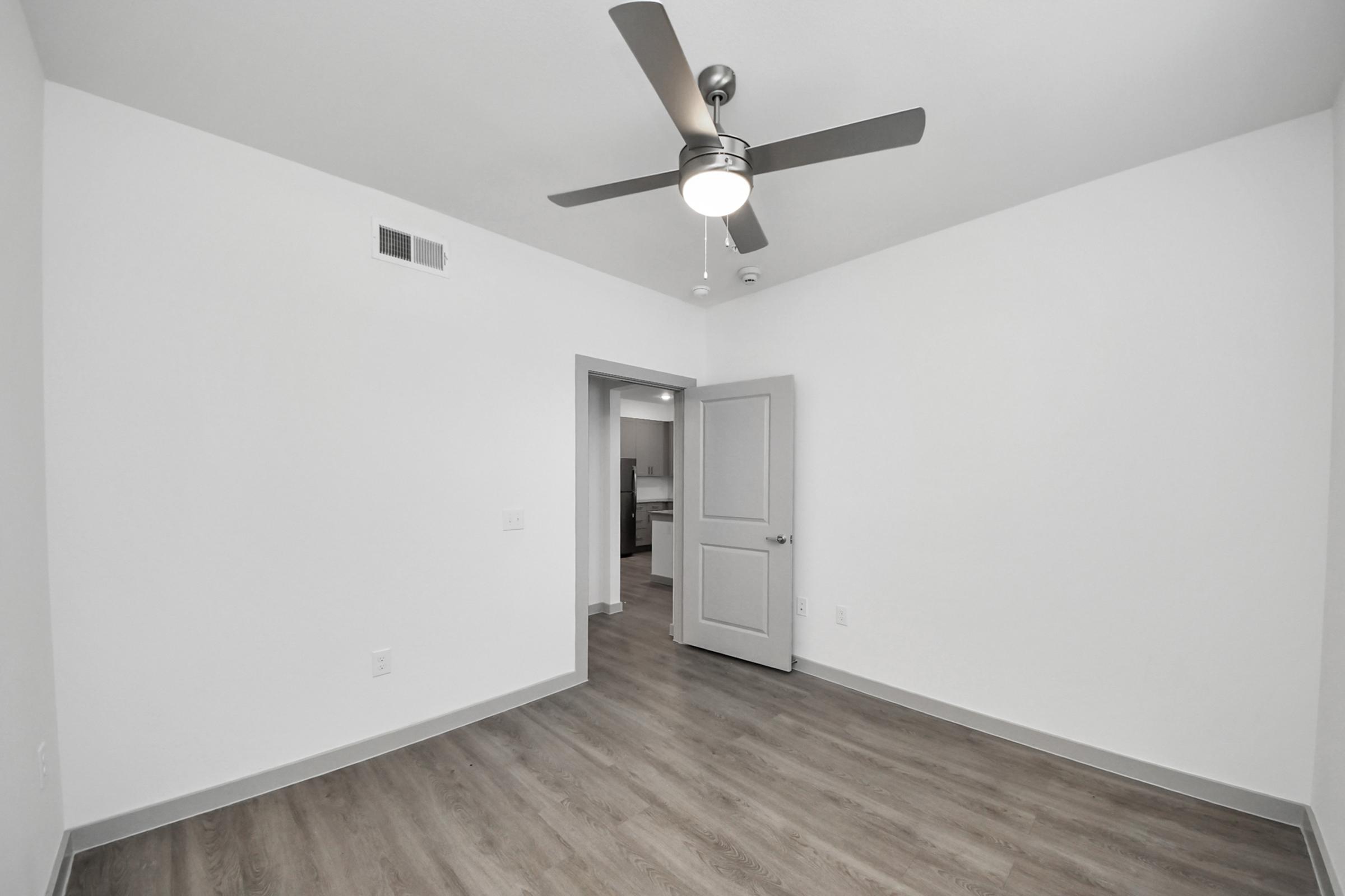 A well-lit, empty room featuring light-colored walls and a modern ceiling fan. The floor is a warm wood tone, and there is a doorway leading to another area. The room is spacious and uncluttered, creating a clean, minimalist aesthetic.