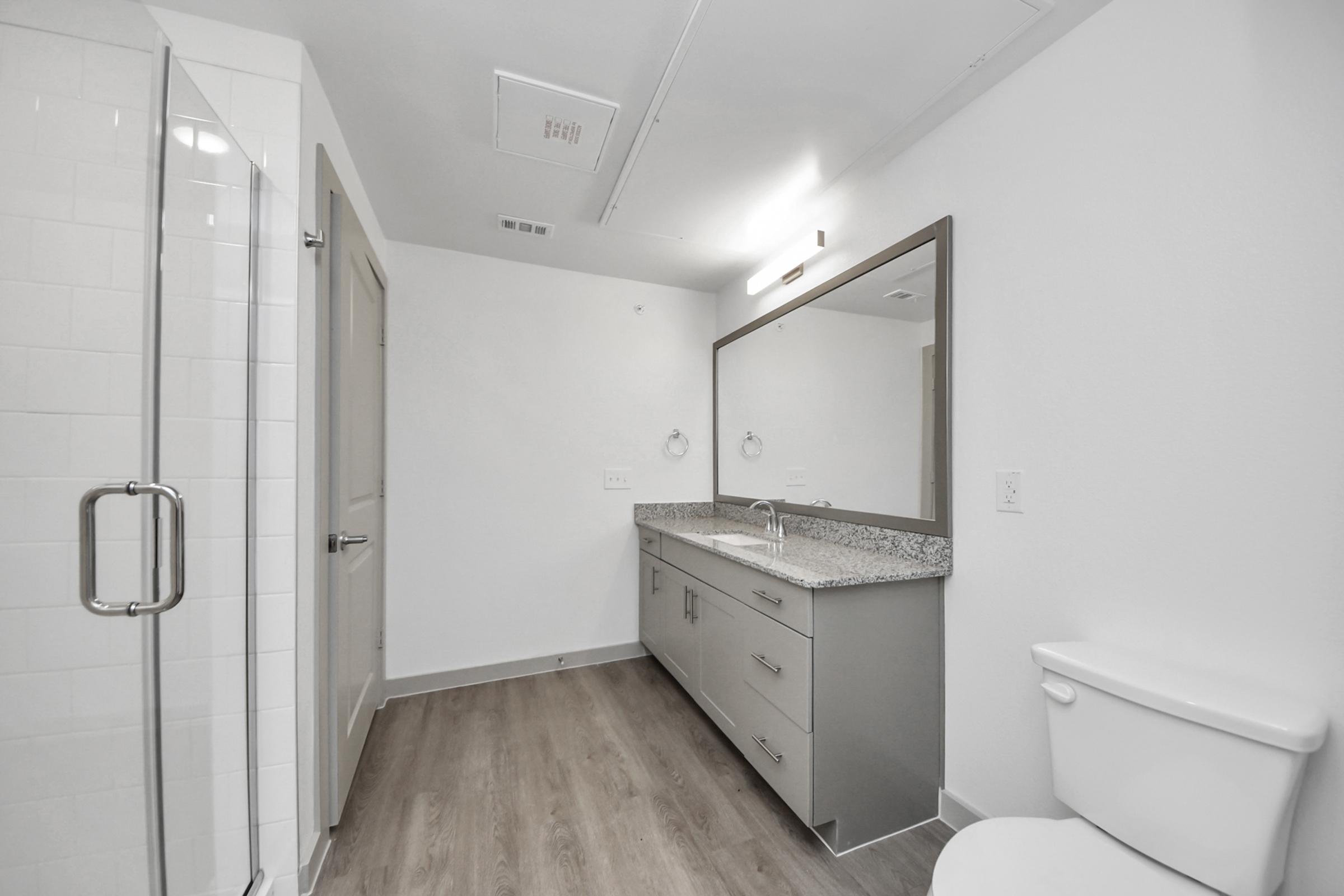 Modern bathroom featuring a walk-in shower with glass doors, a double vanity with a granite countertop, two mirrors, and a toilet. The walls are painted white, and the flooring is light wood. Soft lighting enhances the spacious feel of the room.