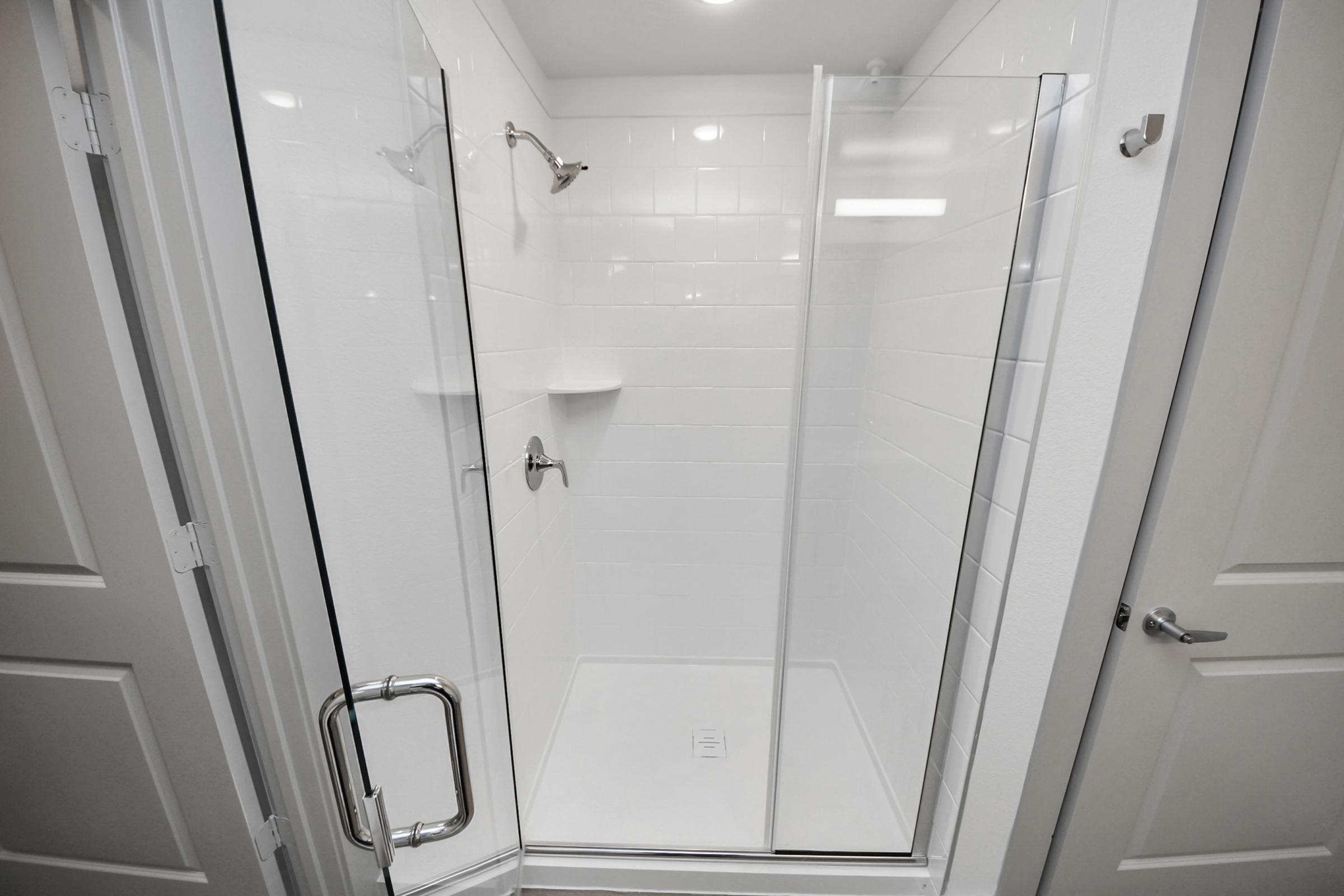 A modern, clean shower enclosure featuring clear glass doors and white tiled walls. It includes a handheld showerhead mounted on the wall and two built-in shelves for toiletries. The design is minimalist and bright, with ample lighting and a sleek handle on the door.