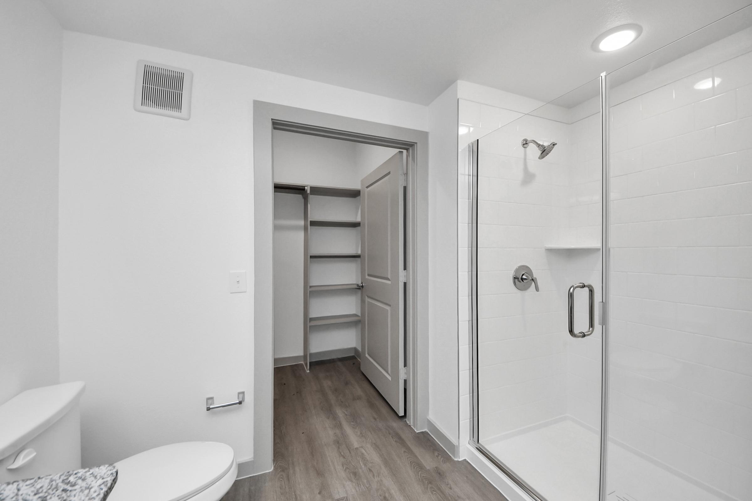 A modern bathroom featuring a glass shower enclosure, toilet, and light-colored walls. There is a door leading to a small closet with shelves, and the flooring is a light wood finish. Bright LED lights illuminate the space.