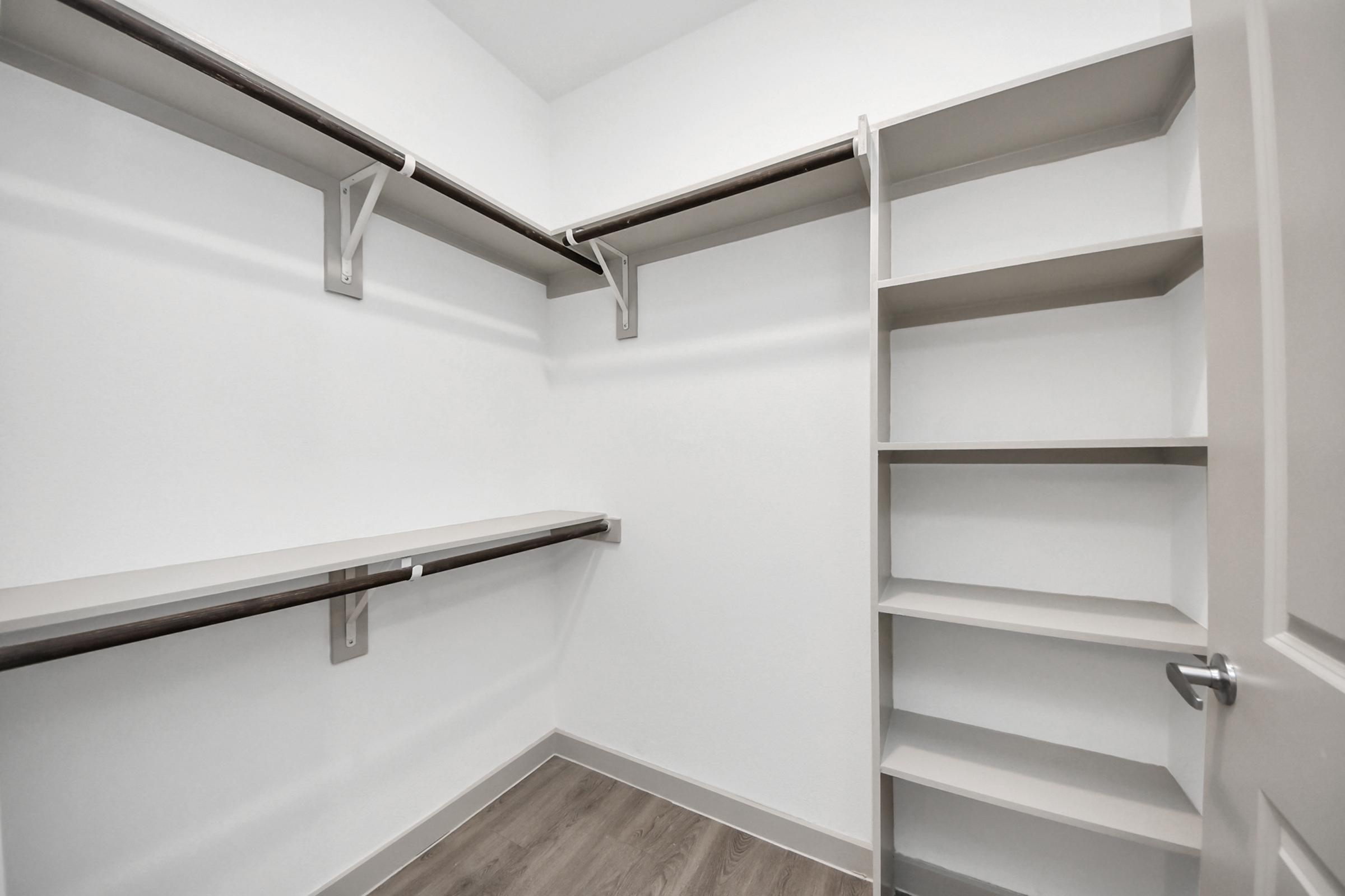A minimalist, empty closet featuring two horizontal shelves and a vertical shelving unit, all against a light-colored wall. The floor has a wood-like finish, giving a clean and organized appearance. The closet door is partially open.