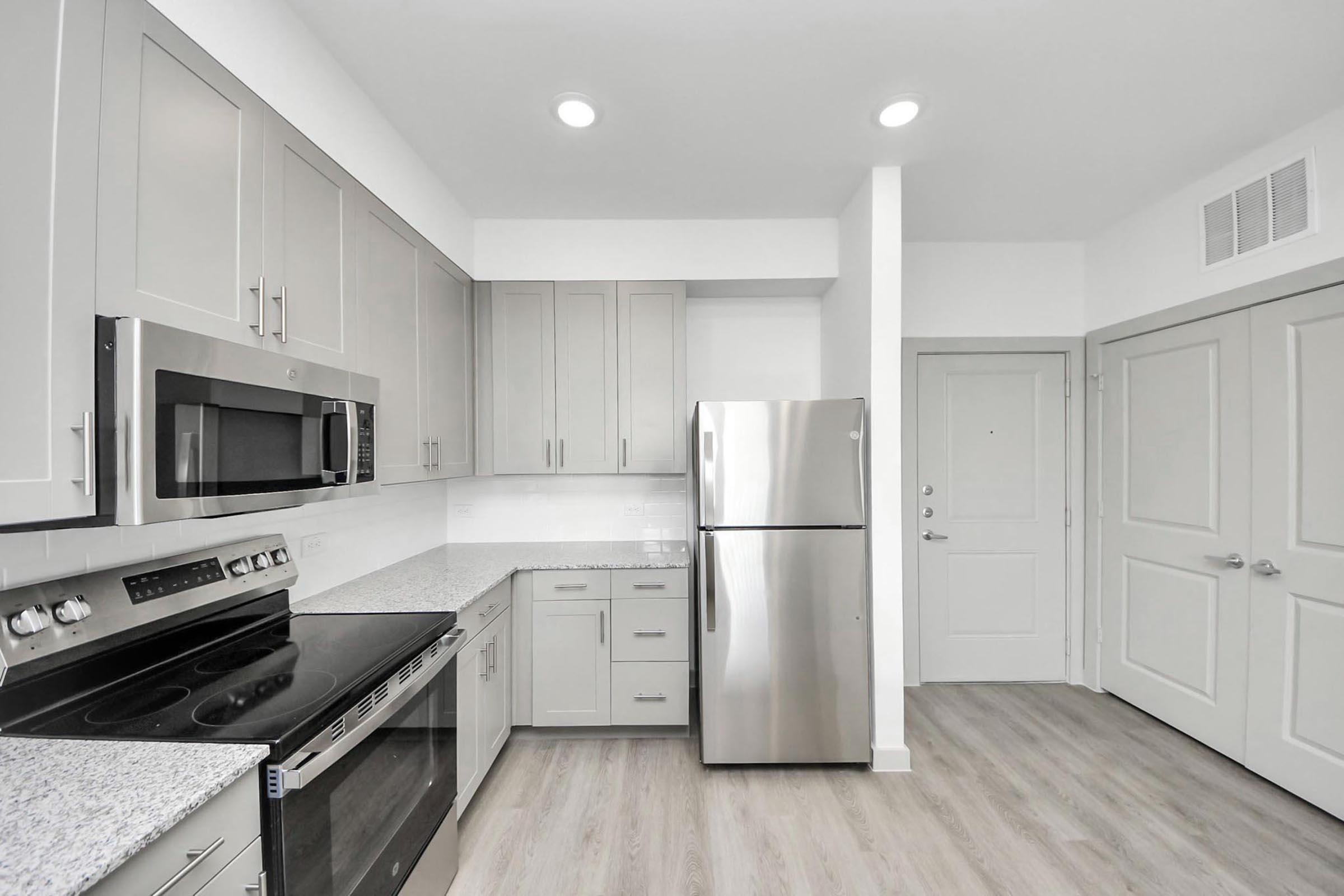 Modern kitchen featuring gray cabinets, stainless steel appliances including a microwave and refrigerator, a granite countertop, and hardwood-style flooring. The space includes an oven and stove, with ample storage and natural light from recessed lighting. A door leads to an adjoining room.