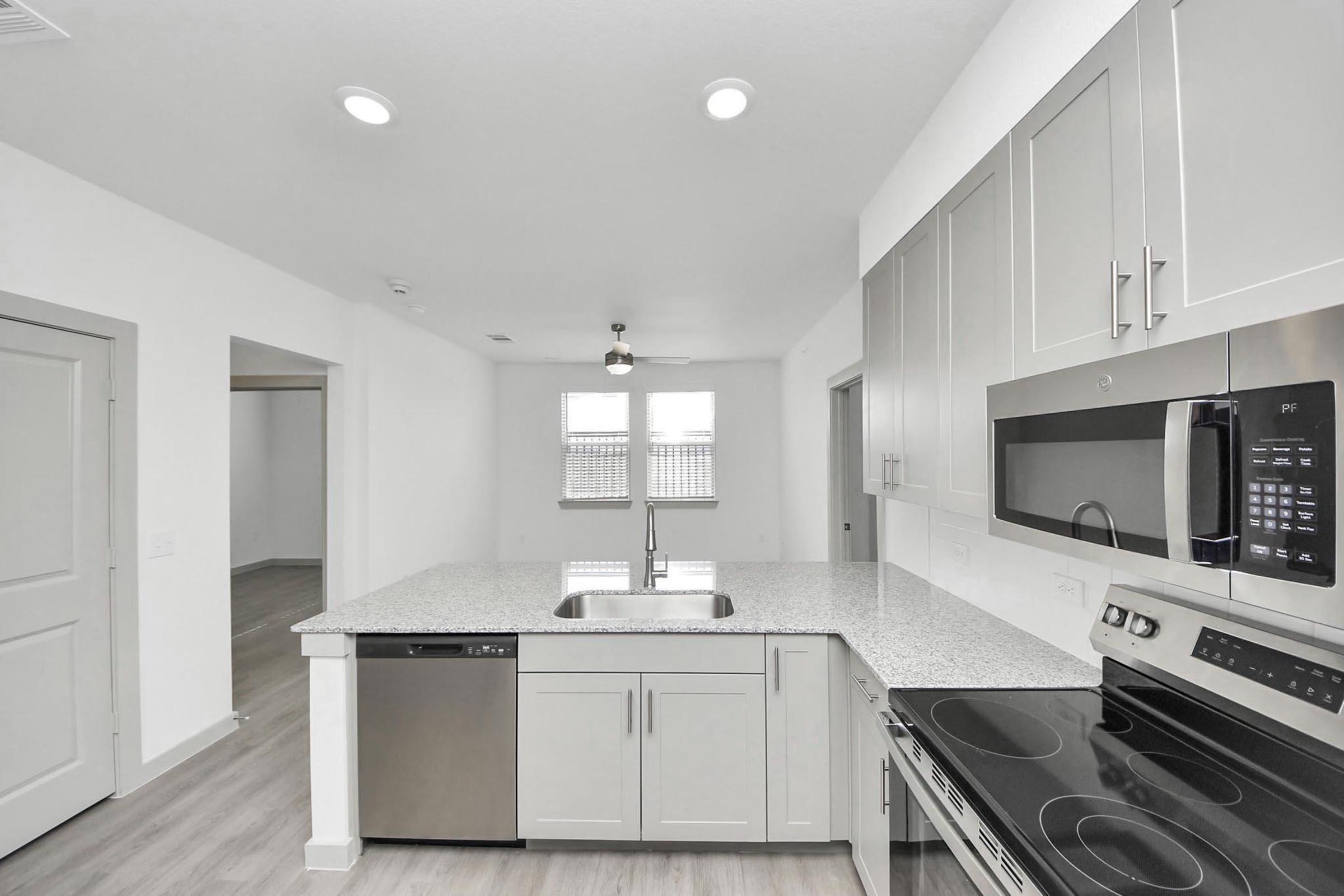 Modern kitchen featuring light gray cabinets, a stainless steel dishwasher, an oven range, and a granite countertop with a sink. The space is bright with recessed lighting and includes a view into an adjacent room with large windows. The flooring is light-colored wood.