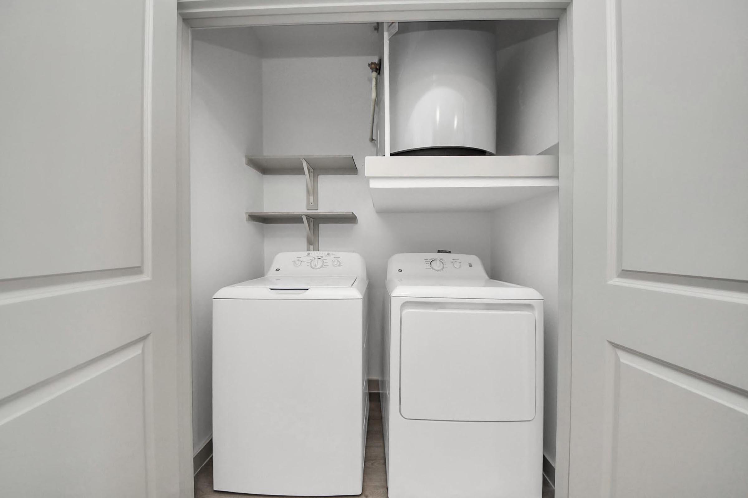 A neatly organized laundry room featuring a white washer and dryer side by side. Above them, there is a shelf with additional space for storage. The walls are painted light gray, and the area is well-lit, creating a clean and modern look.