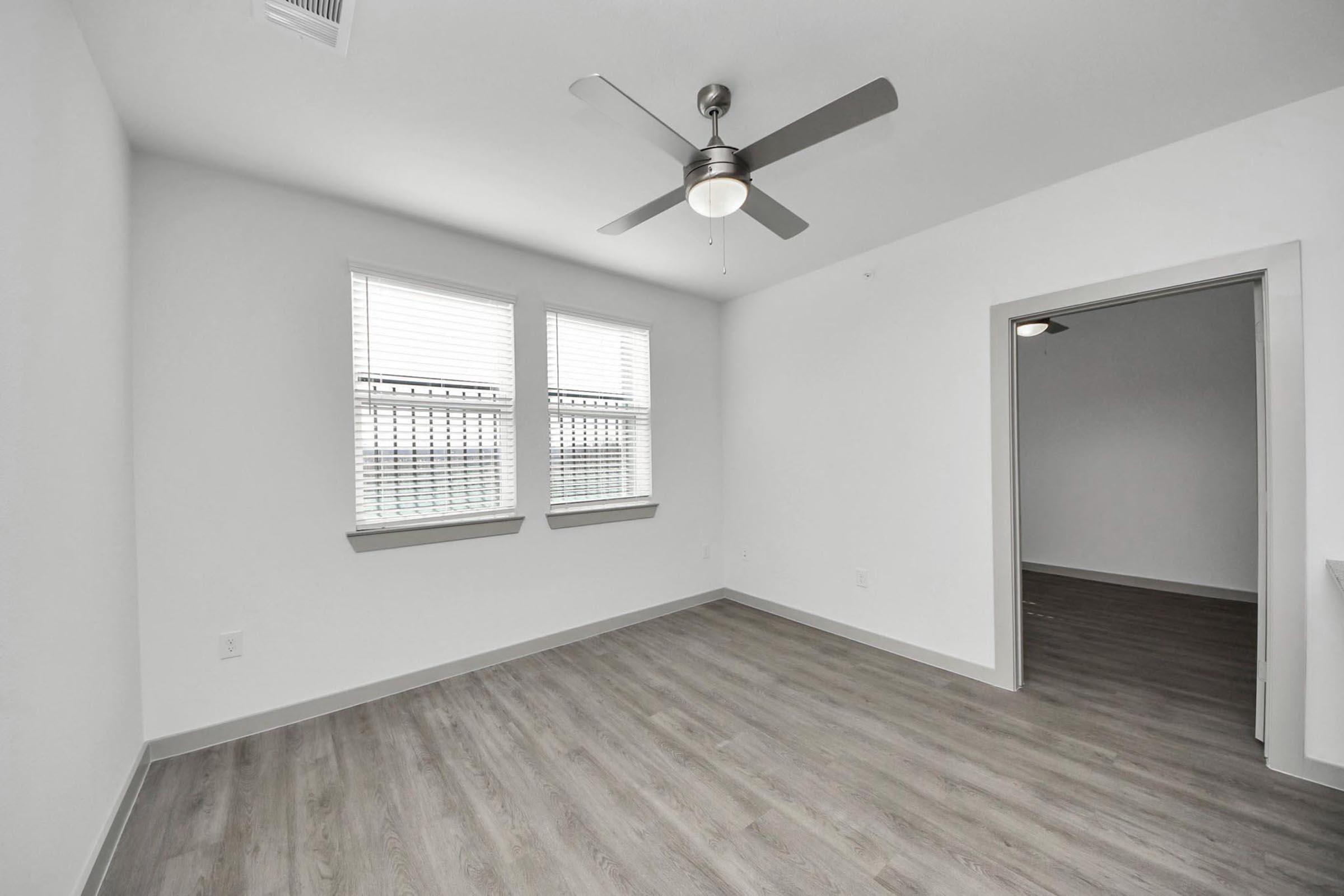 A spacious, minimalist room with light gray walls and wood-like flooring. It features two windows with blinds allowing natural light, a modern ceiling fan, and an open doorway leading to another room. The overall aesthetic is clean and uncluttered, suitable for various interior design styles.