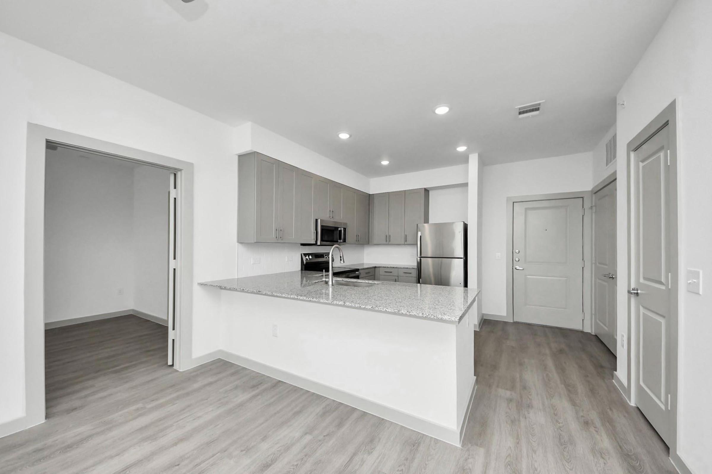 A modern kitchen featuring light wood flooring, gray cabinets, stainless steel appliances, and a central island with a granite countertop. There are bright overhead lights and an open layout leading to a living space, with a partially visible door on the left.