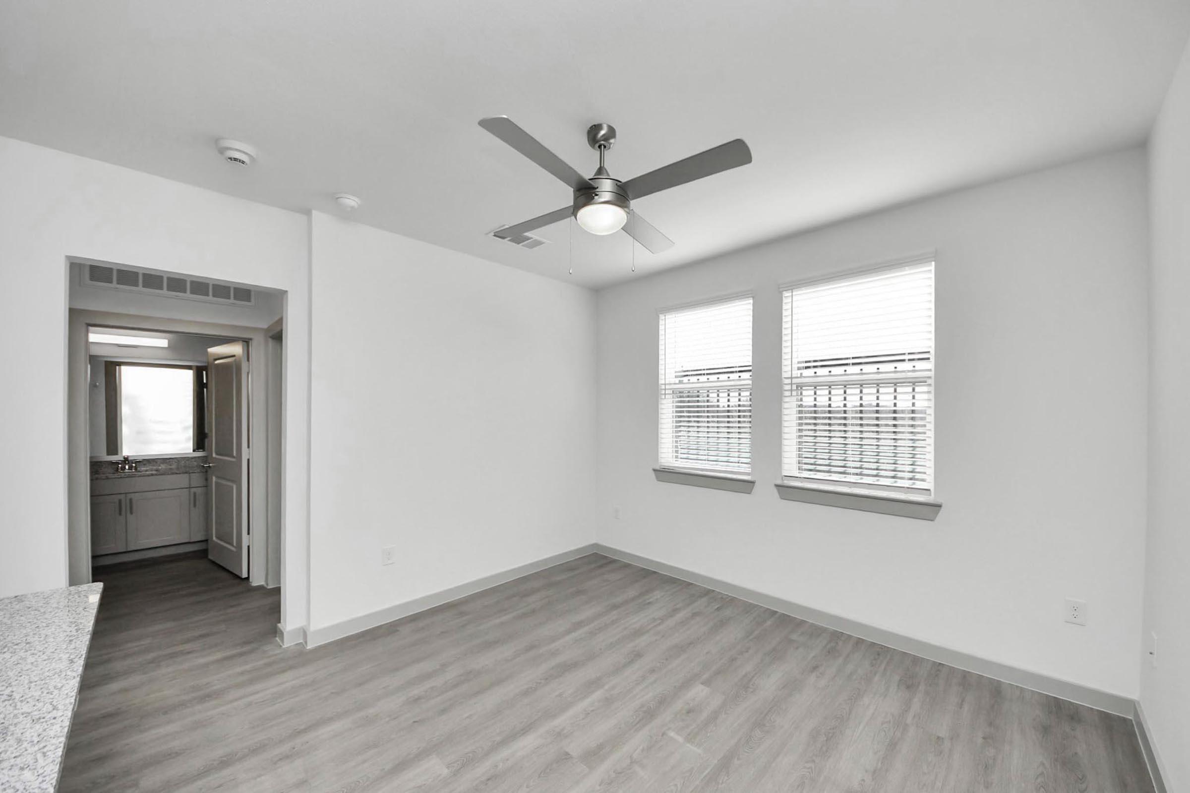 A bright and empty living room featuring light-colored walls, large windows with blinds, and a ceiling fan. An adjoining doorway leads to a bathroom area with modern fixtures. The flooring is a light wood finish, creating a spacious and open atmosphere.