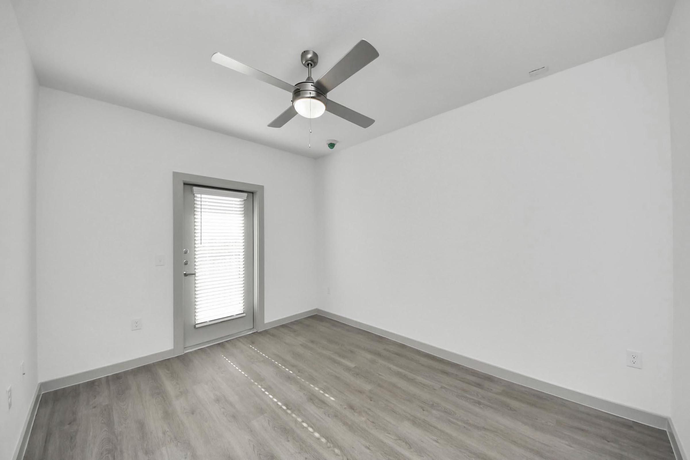 Interior view of a minimalist room featuring light gray walls, a ceiling fan, and a window with blinds. The floor is a light wood laminate. The room is empty, creating a spacious and airy feel, and there is a door leading outside.