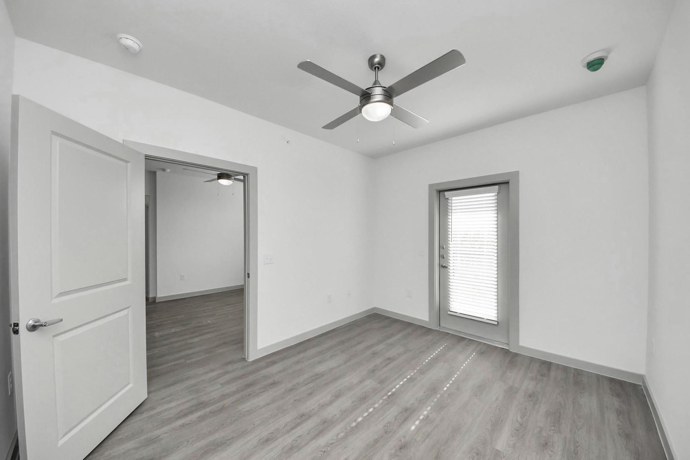 A spacious, modern room with light gray walls and a ceiling fan. Two doorways lead to adjacent areas, and a large window with blinds allows natural light to enter. The flooring is light wood, enhancing the airy feel of the space.