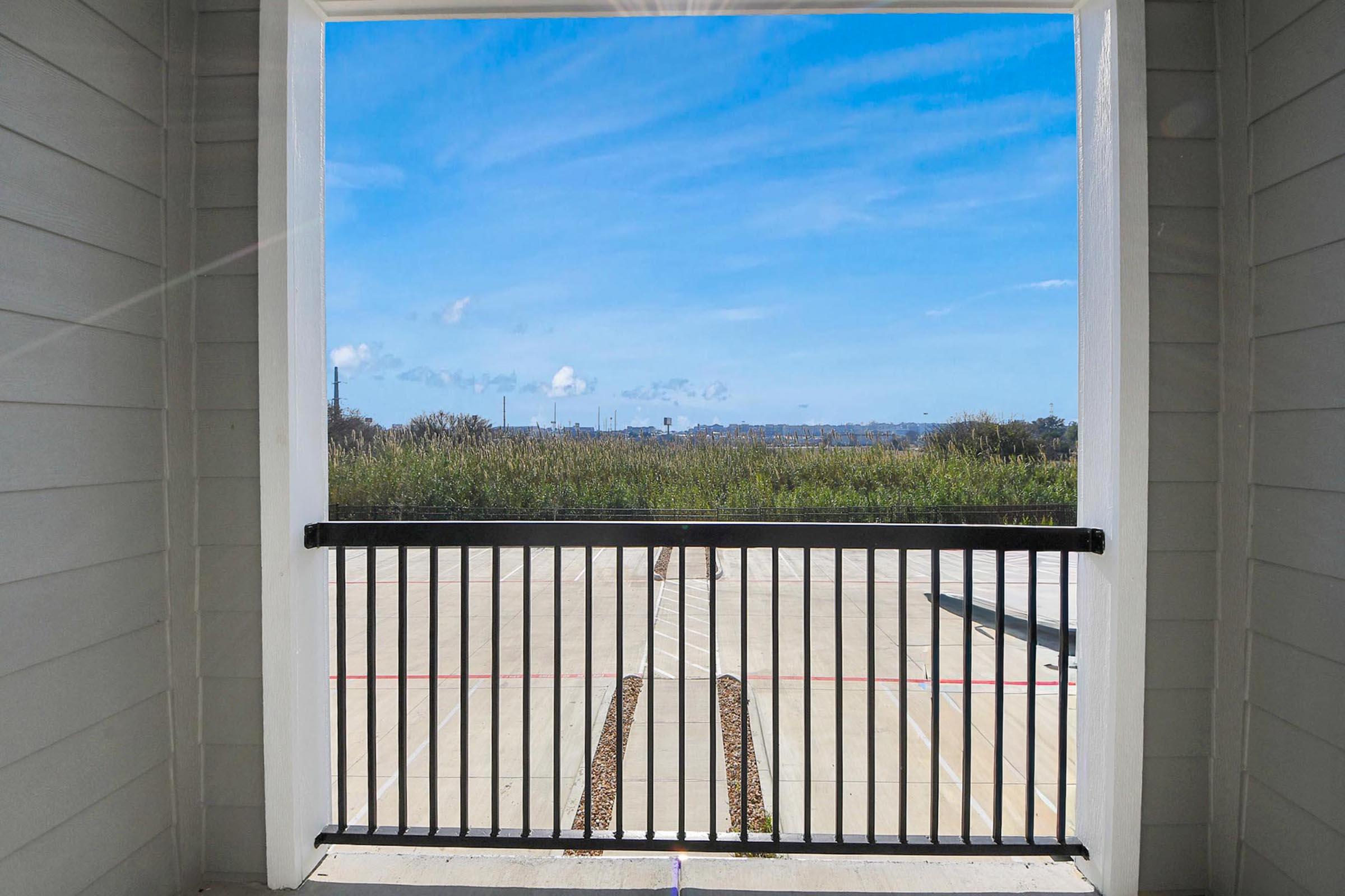 A view from a balcony featuring a black railing, overlooking a wide expanse of greenery and a clear blue sky with scattered clouds. The scene includes a paved area below, hinting at a tranquil outdoor space.