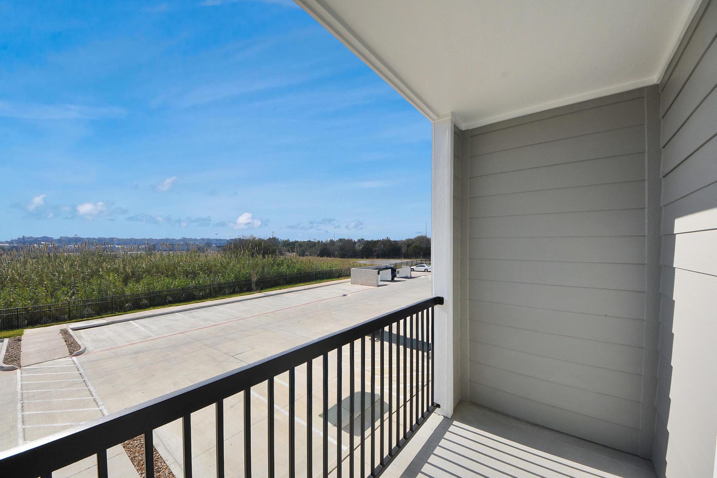 A view from a balcony showing a clear blue sky, with a few clouds and a scenic landscape featuring green vegetation. Below is a paved area with parking spaces and a road in the distance, creating a peaceful outdoor atmosphere.