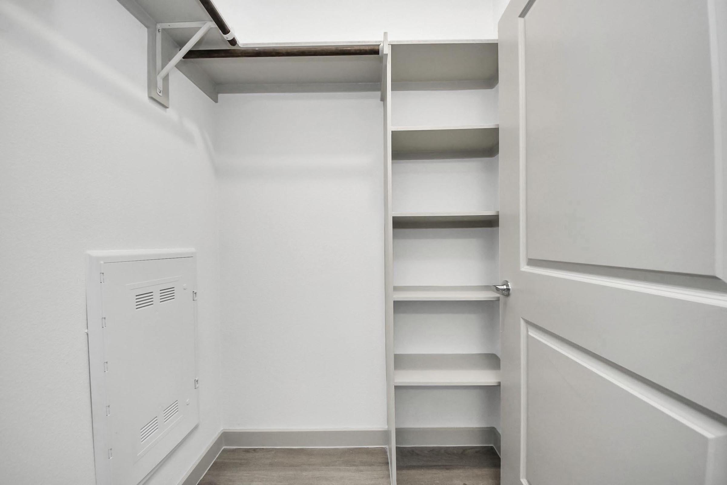 A clean, minimalist storage closet featuring white walls and a built-in shelving unit. The closet has a door on the right, and the flooring is a light wood finish. An electrical panel is visible on the left wall, with ample space for organization.