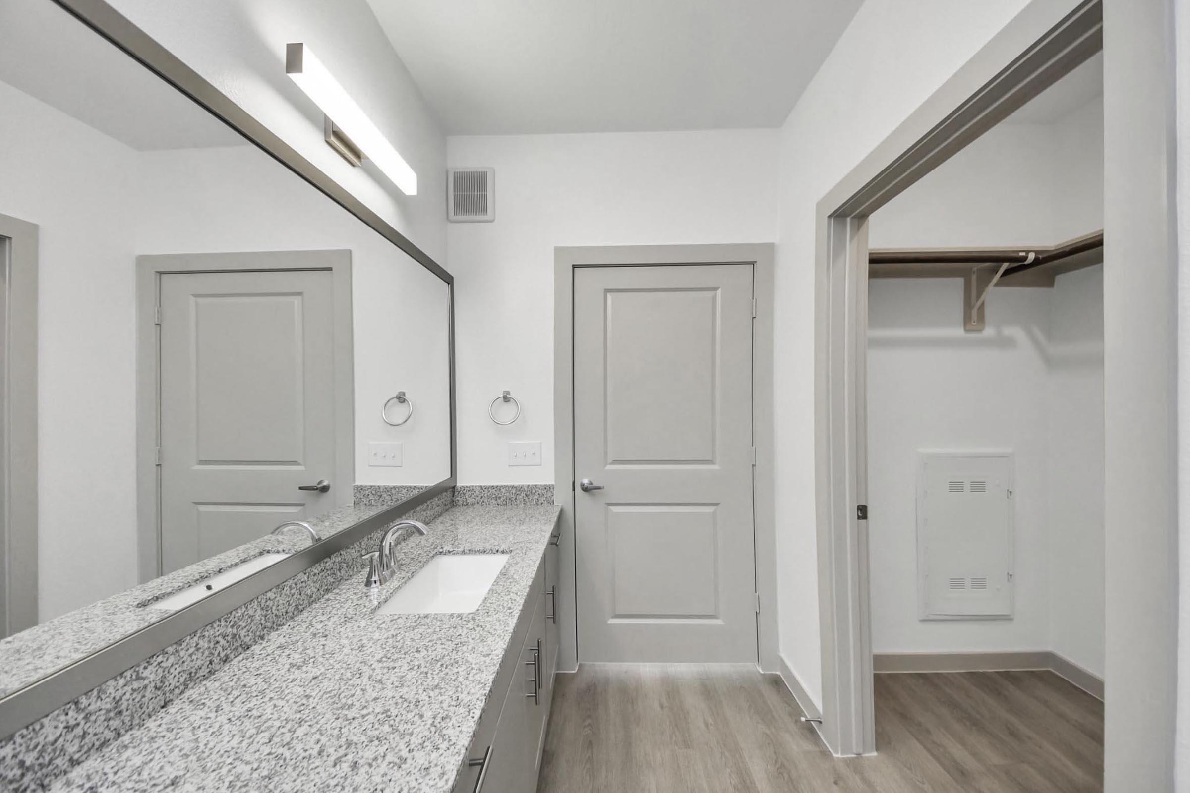 A modern bathroom featuring a double sink vanity with a gray granite countertop, large wall-mounted mirror, gray cabinetry, and minimalistic fixtures. The space has light-colored walls and flooring, with a door leading to a closet area on the right. Bright lighting enhances the clean and contemporary design.