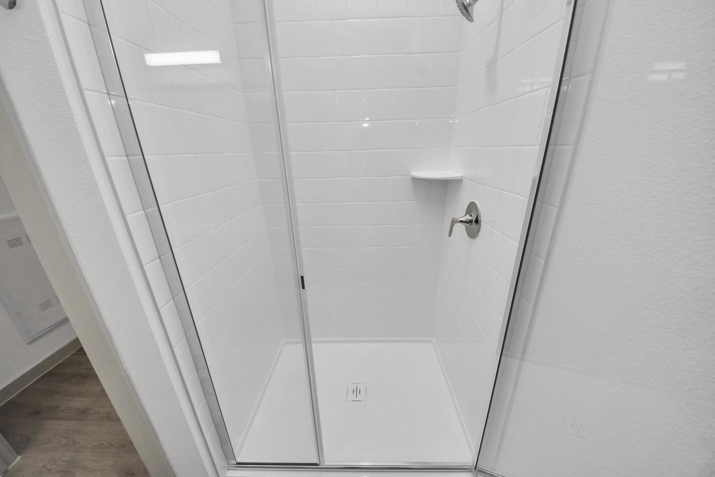 A clean and modern shower with glass doors, featuring white tile walls and a built-in shelf. The floor is tiled, and there is a chrome showerhead mounted on the wall, creating a sleek and minimalist design. The overall ambiance is bright and fresh.