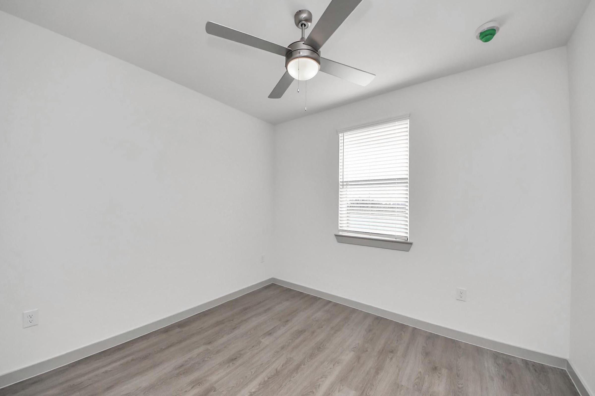 Empty room with light gray walls and wooden flooring. A ceiling fan with a light fixture is mounted in the center, and there is one window with blinds on the left side, letting in natural light. The space is minimally decorated, featuring clean lines and a bright, airy atmosphere.