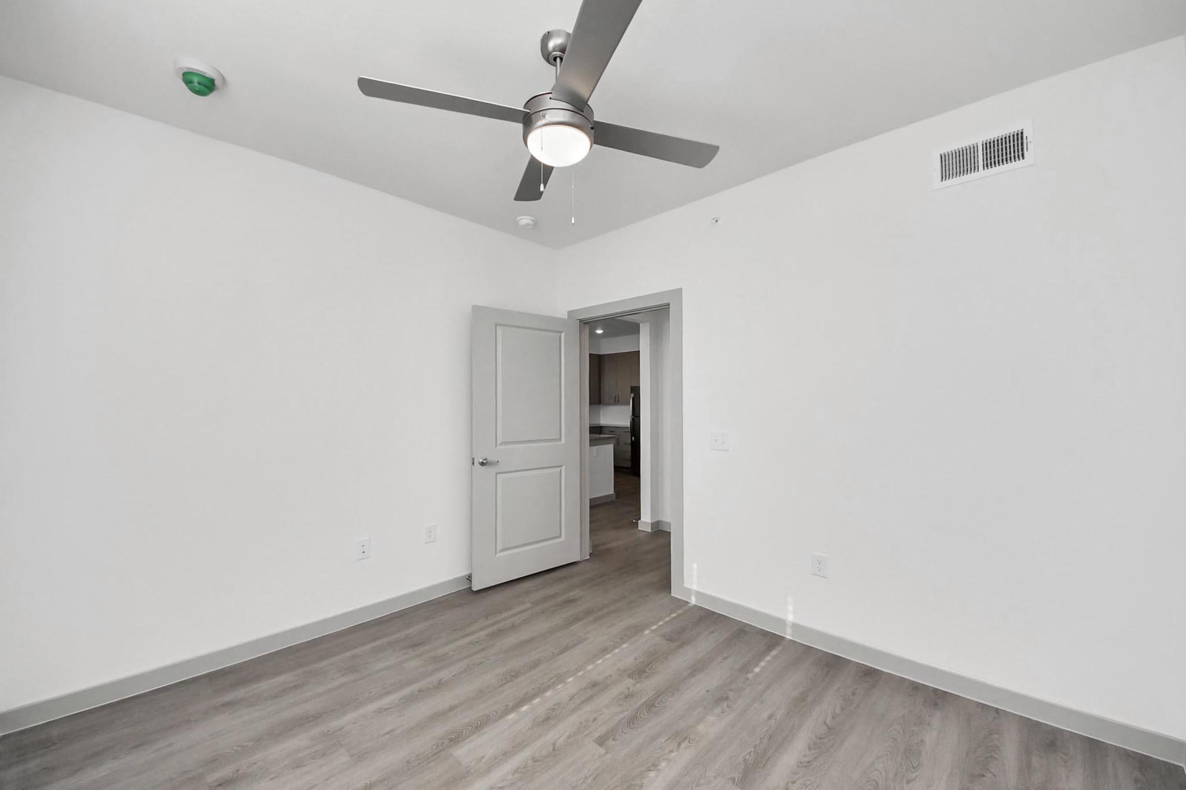 A bright, empty room with light gray walls and a ceiling fan. There is a partially open door leading to another room, and the floor is covered in light wood-like laminate. A smoke detector is visible on the ceiling, and the overall ambiance is clean and modern.
